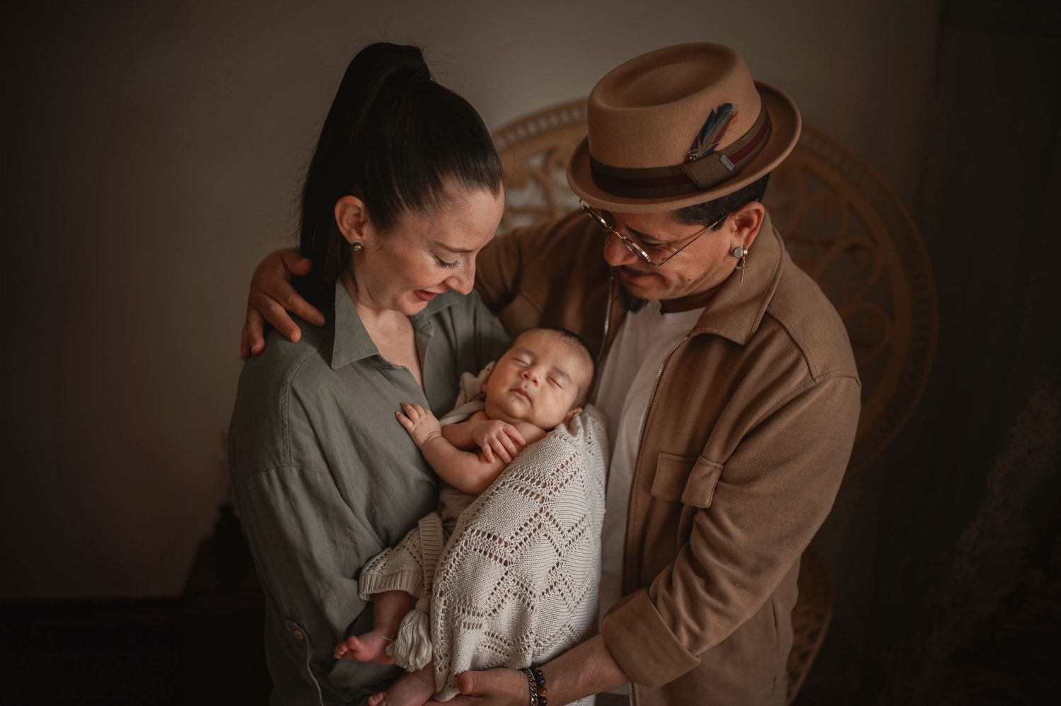 Intimate family portrait with sleeping infant wrapped in white knit blanket in warm vintage tones.