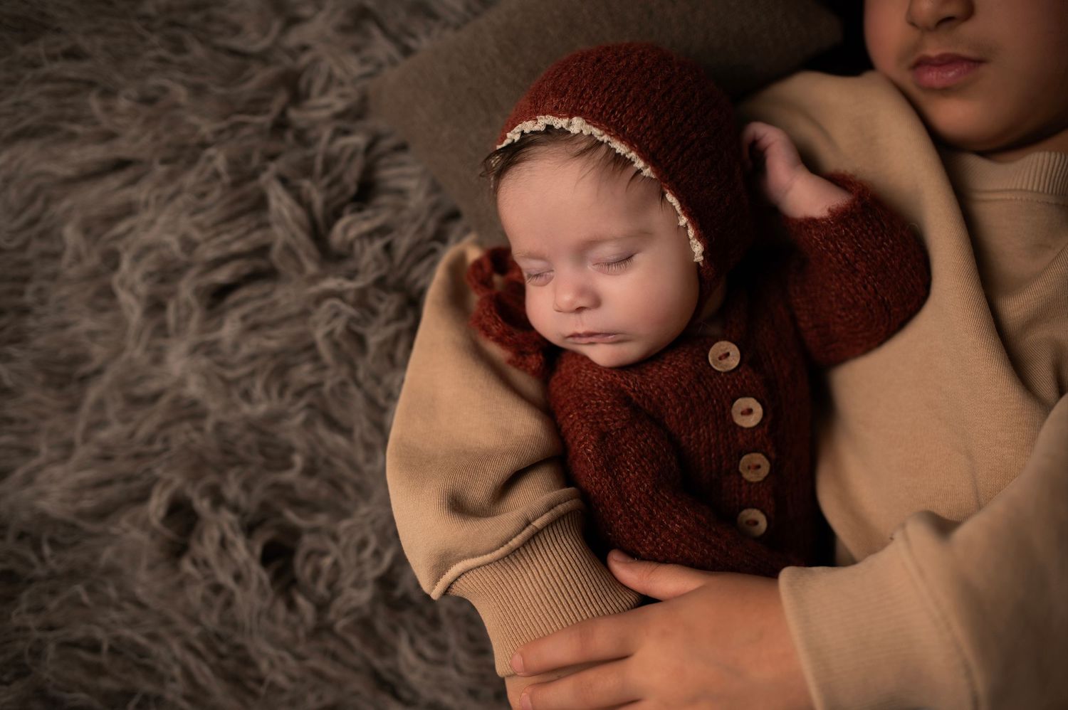 Sleeping baby in burgundy cardigan nestled in tan blanket on textured surface.