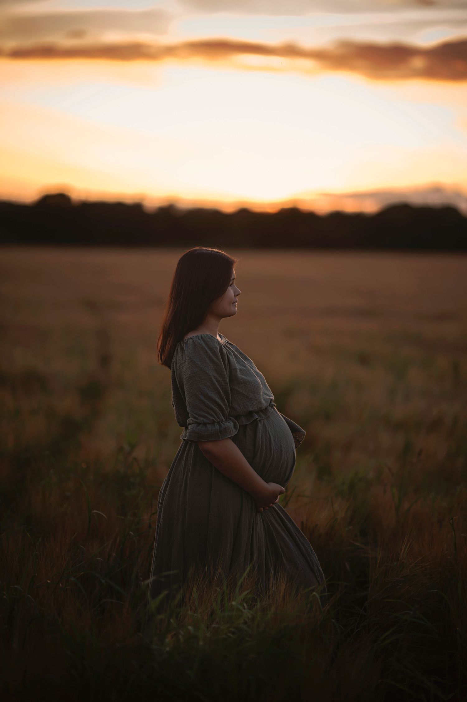 Silhouette of pregnant woman in dress standing in field at sunset.