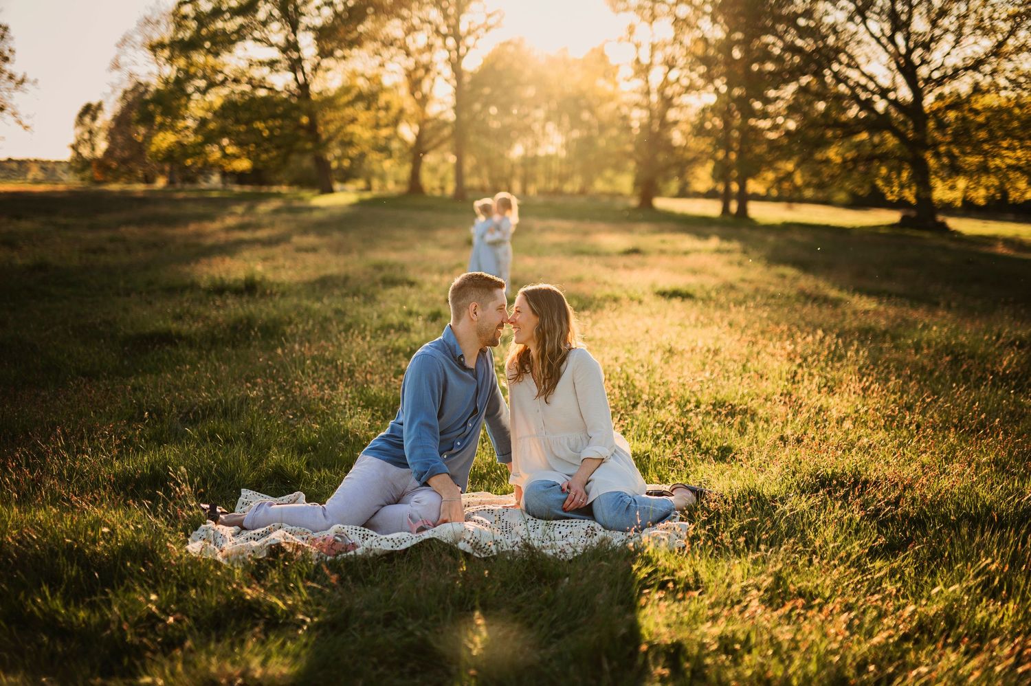 Familjefotografering i Skåne med par på filt i solig äng med barn i bakgrunden.