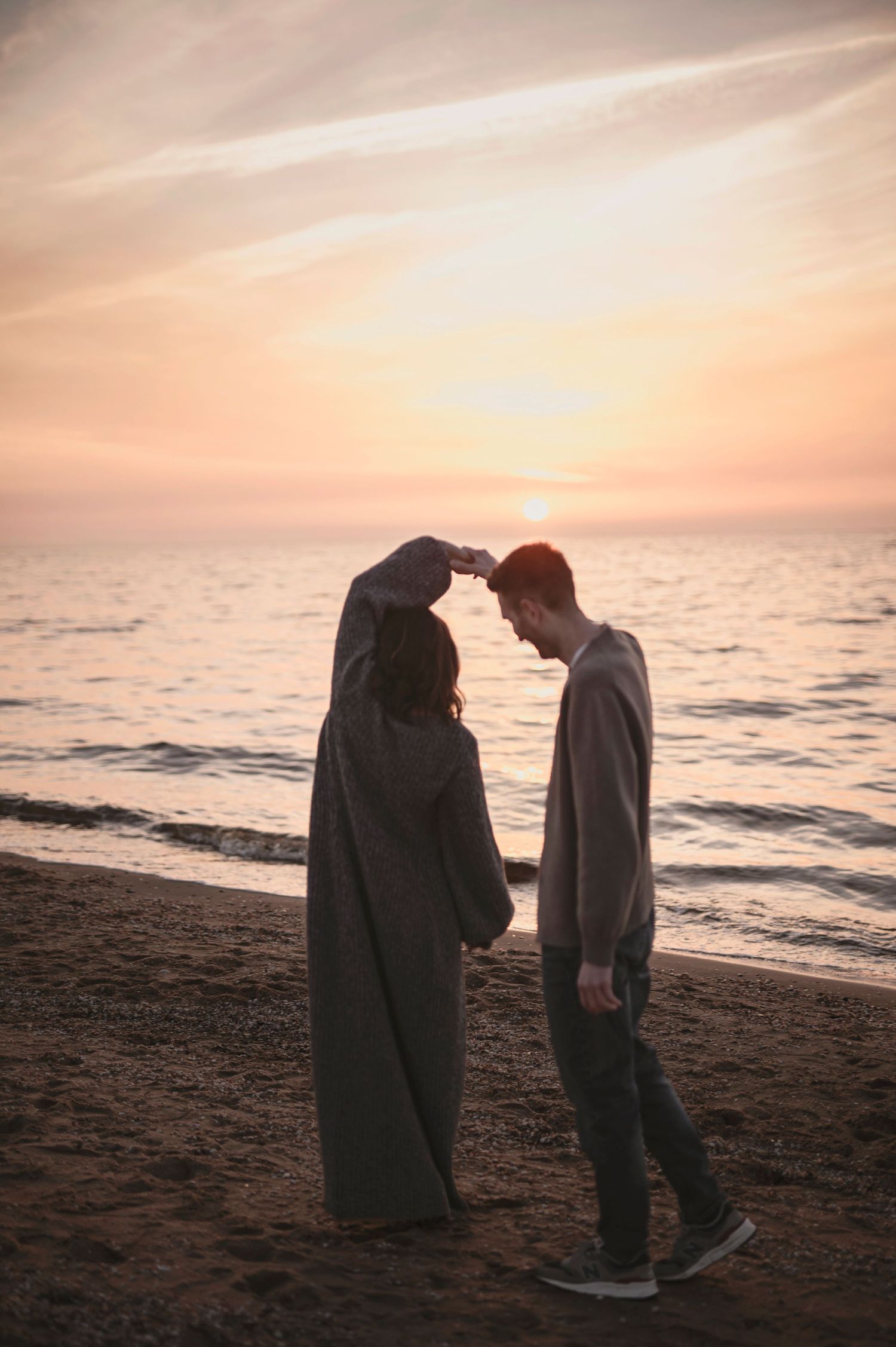 A couple stands together on a beach at sunset, silhouetted against the golden sky and ocean waves.