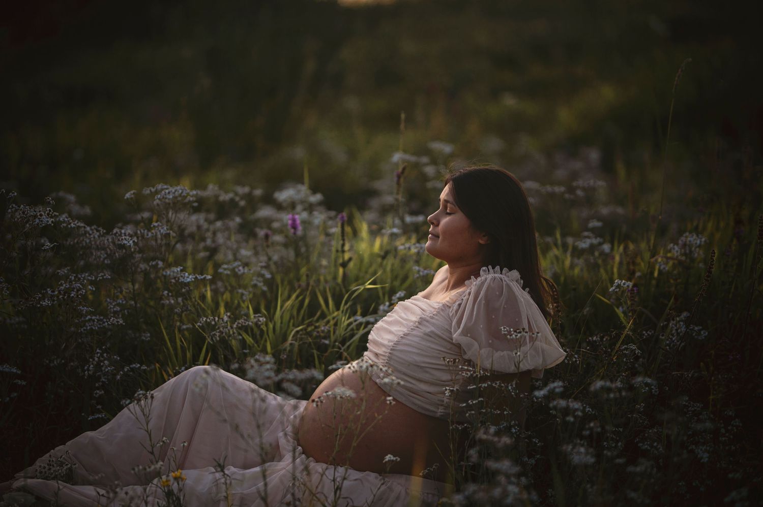 A dreamy maternity photo among wildflowers at dusk in a white off-shoulder dress.