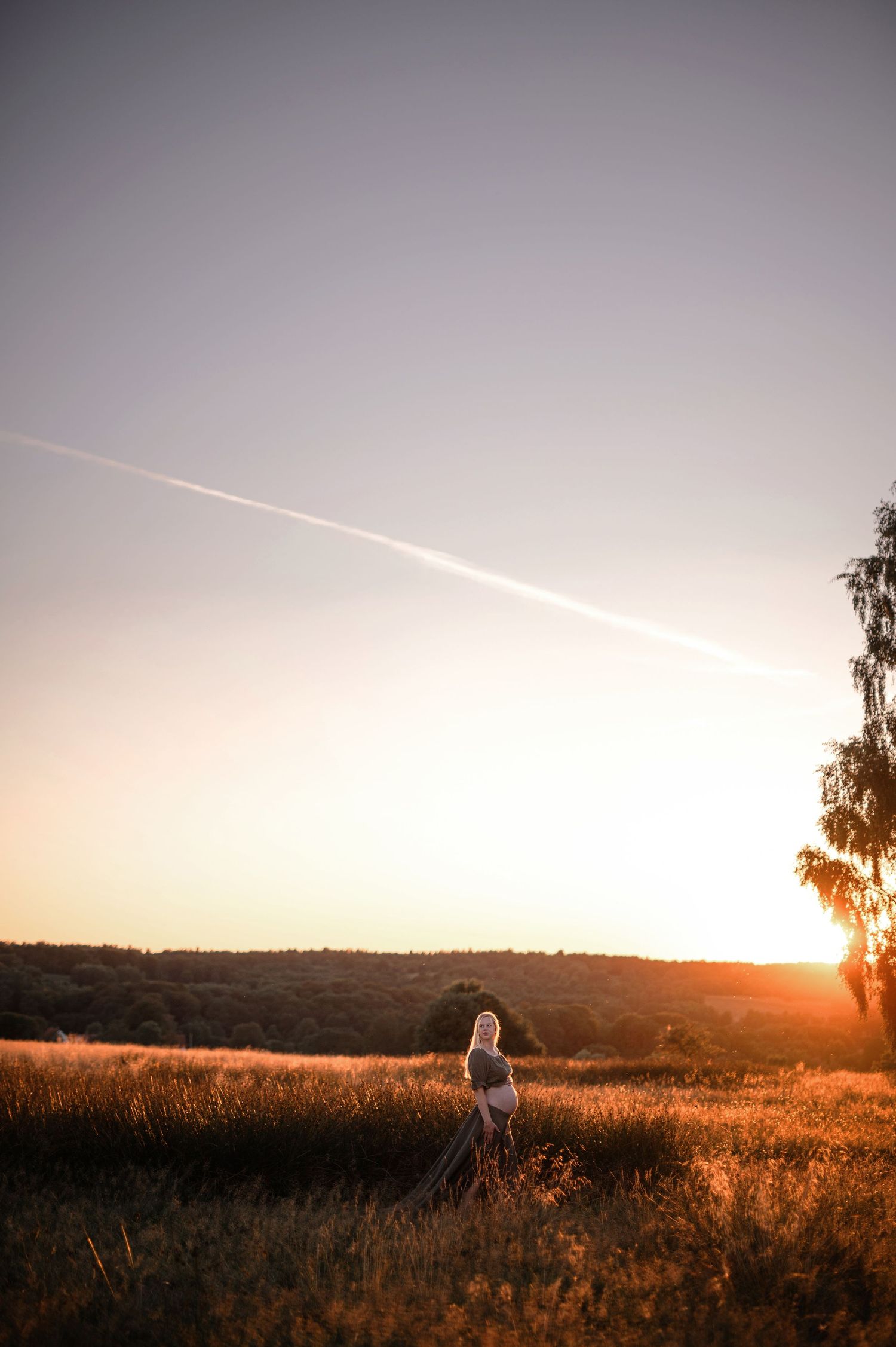 Woman standing in golden field at sunset with airplane contrail across clear sky and trees silhouetted on horizon.