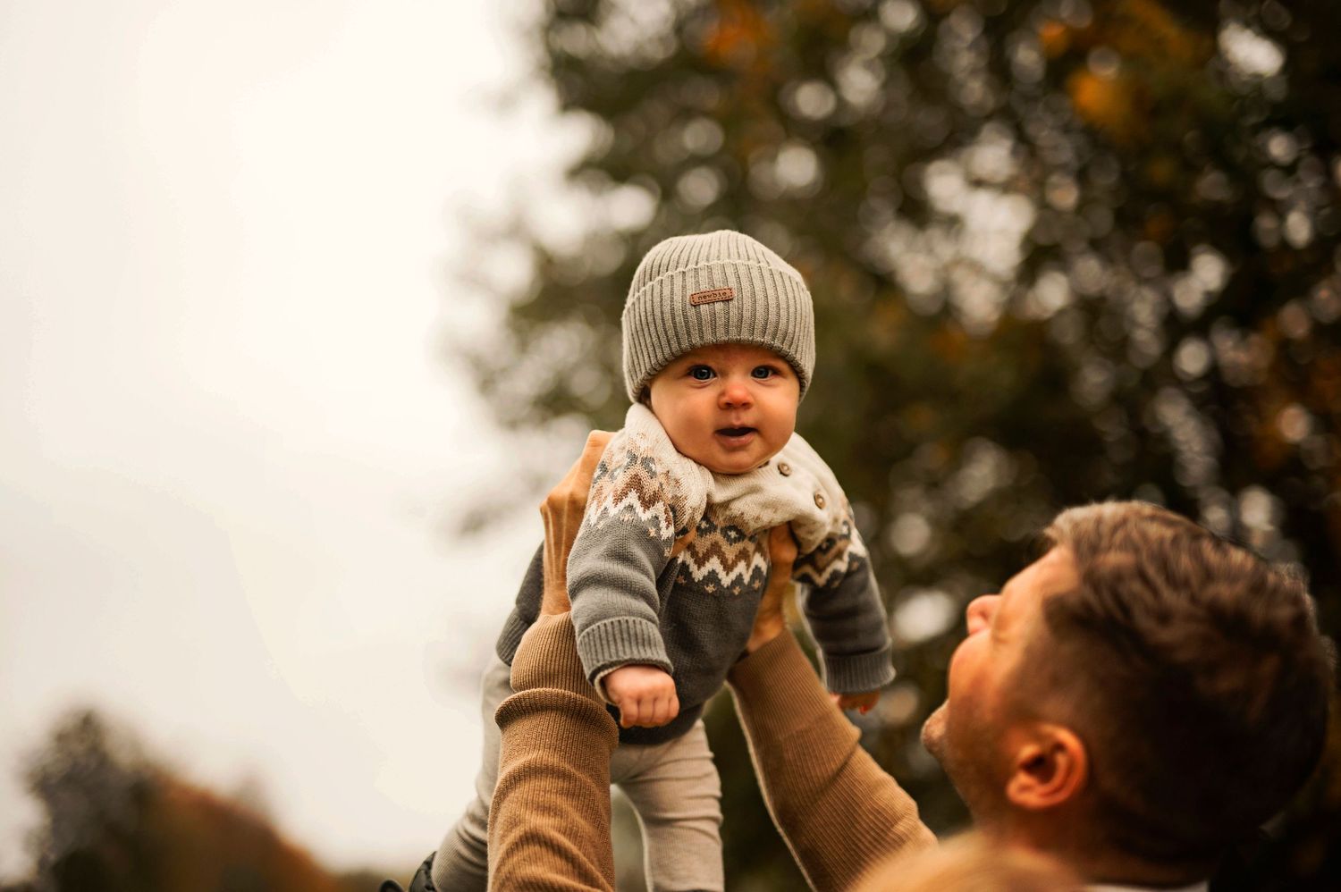 A baby wearing a knit hat is lifted high in the autumn air.