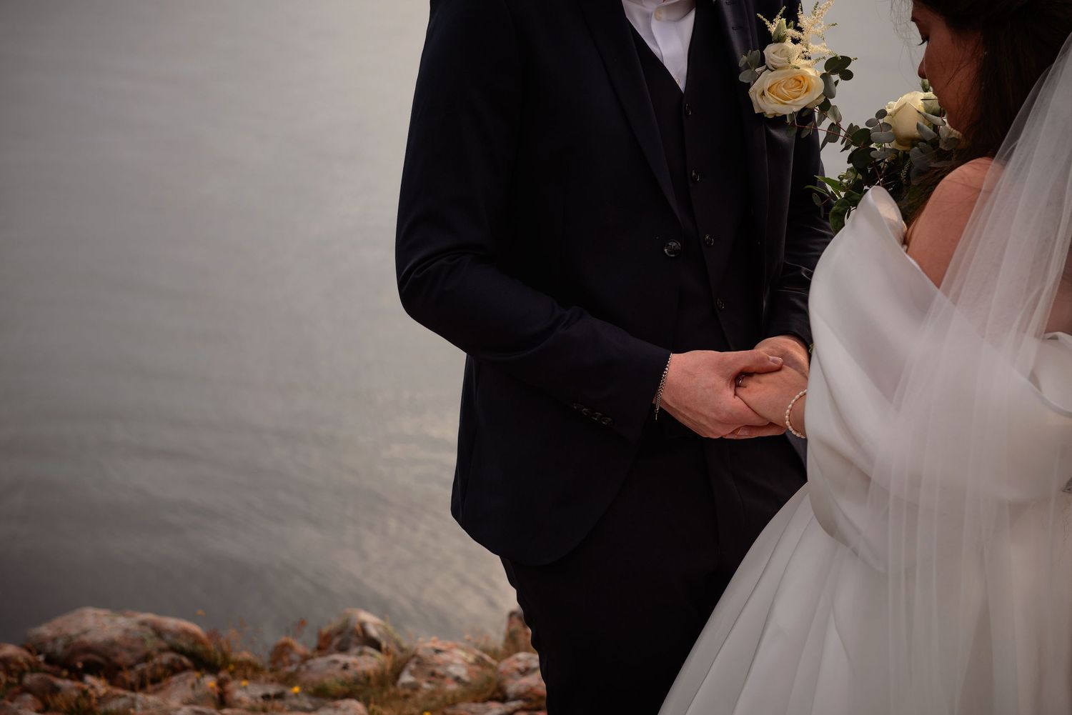 Bride and groom holding hands during outdoor wedding ceremony by a lake, bride holding white floral bouquet.