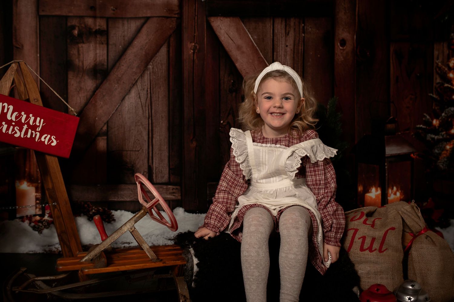 Young girl in holiday outfit sits smiling by Christmas sled and decorations in rustic barn setting.