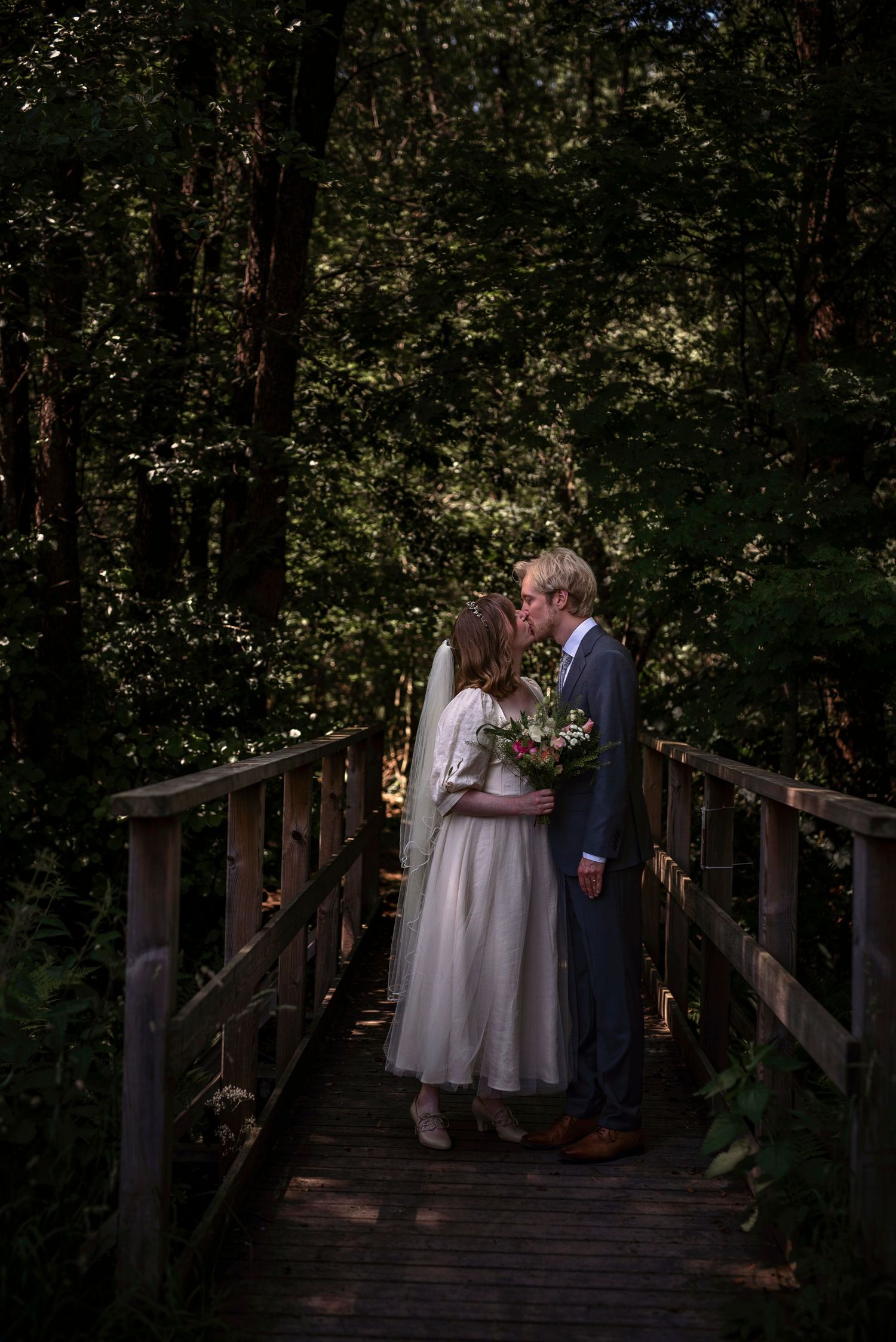A bride and groom share a kiss on a wooden bridge surrounded by lush green forest trees on their wedding day.
