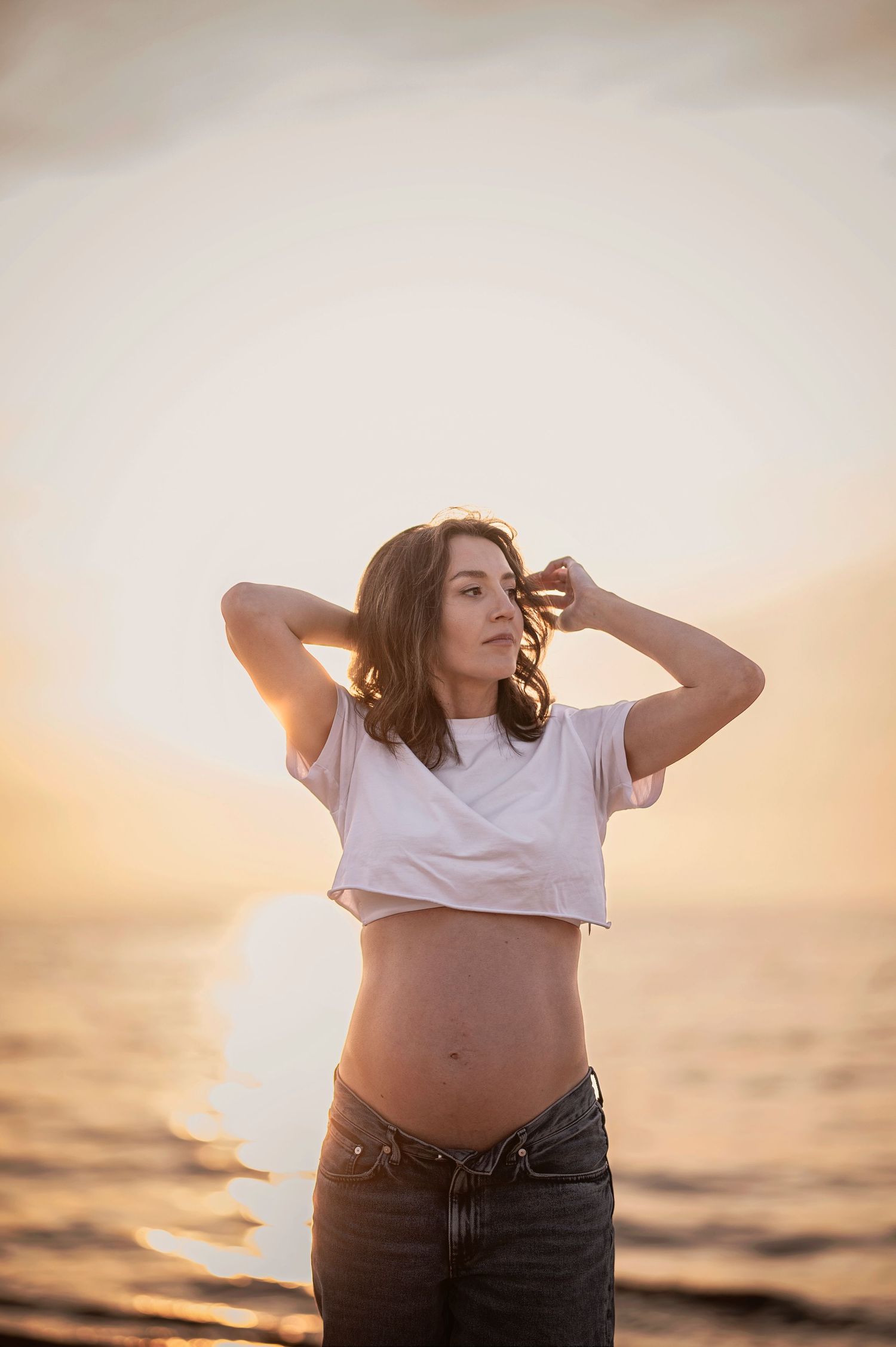 Pregnant woman in crop top and jeans poses against golden sunset over ocean.