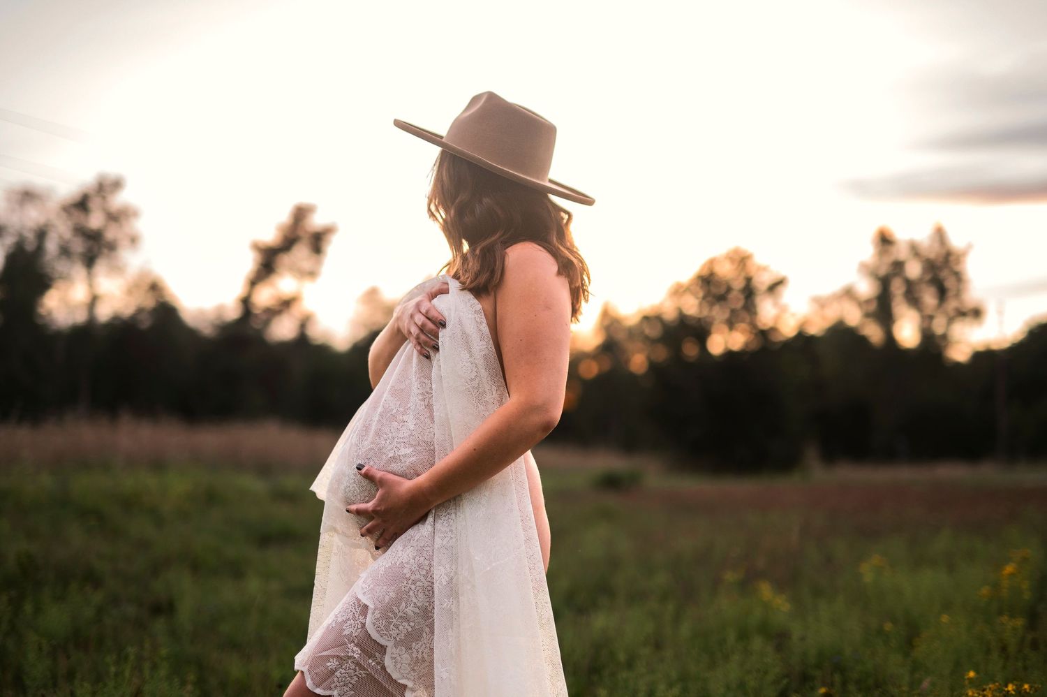Gravidfotografering i Skåne med blivande mamma i vit spetsklänning i rustikt fält i kvällsljus.