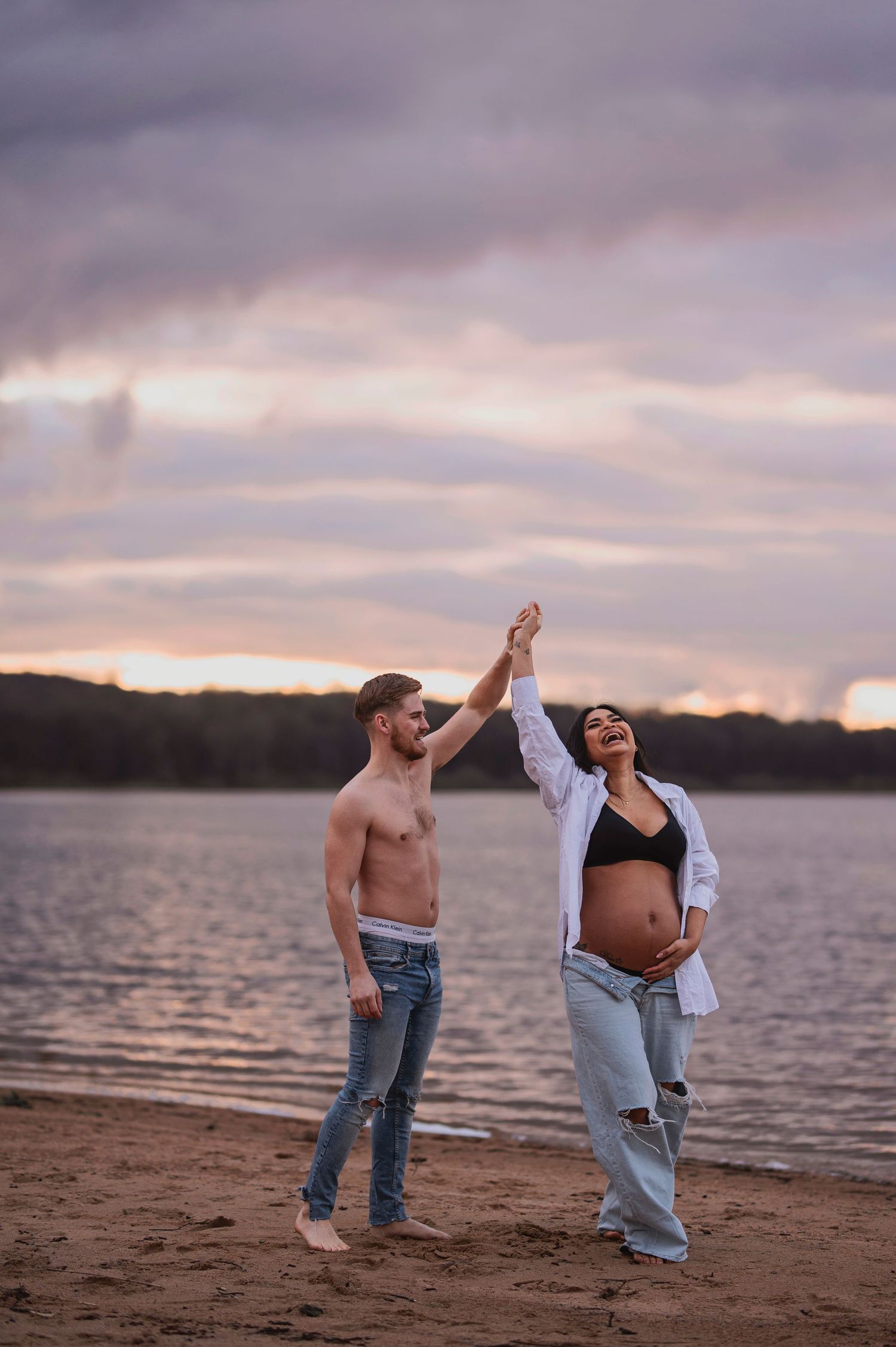 A couple holds hands high in celebration on a lakeside beach during sunset.