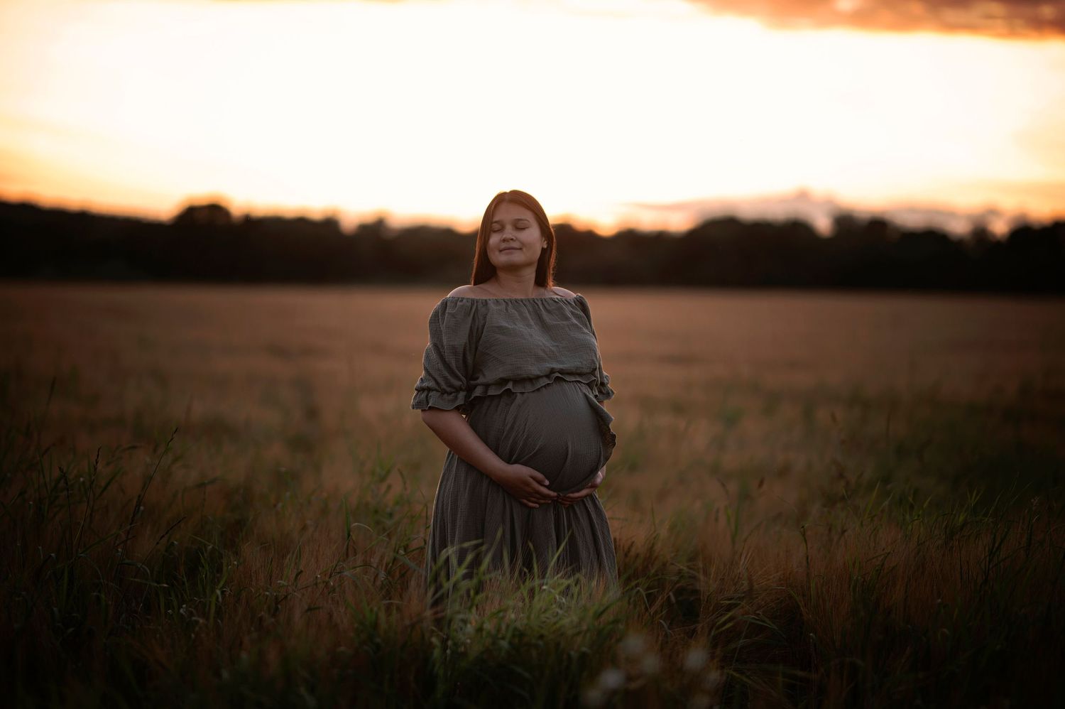A pregnant silhouette stands in a grassy field during a dramatic sunset.