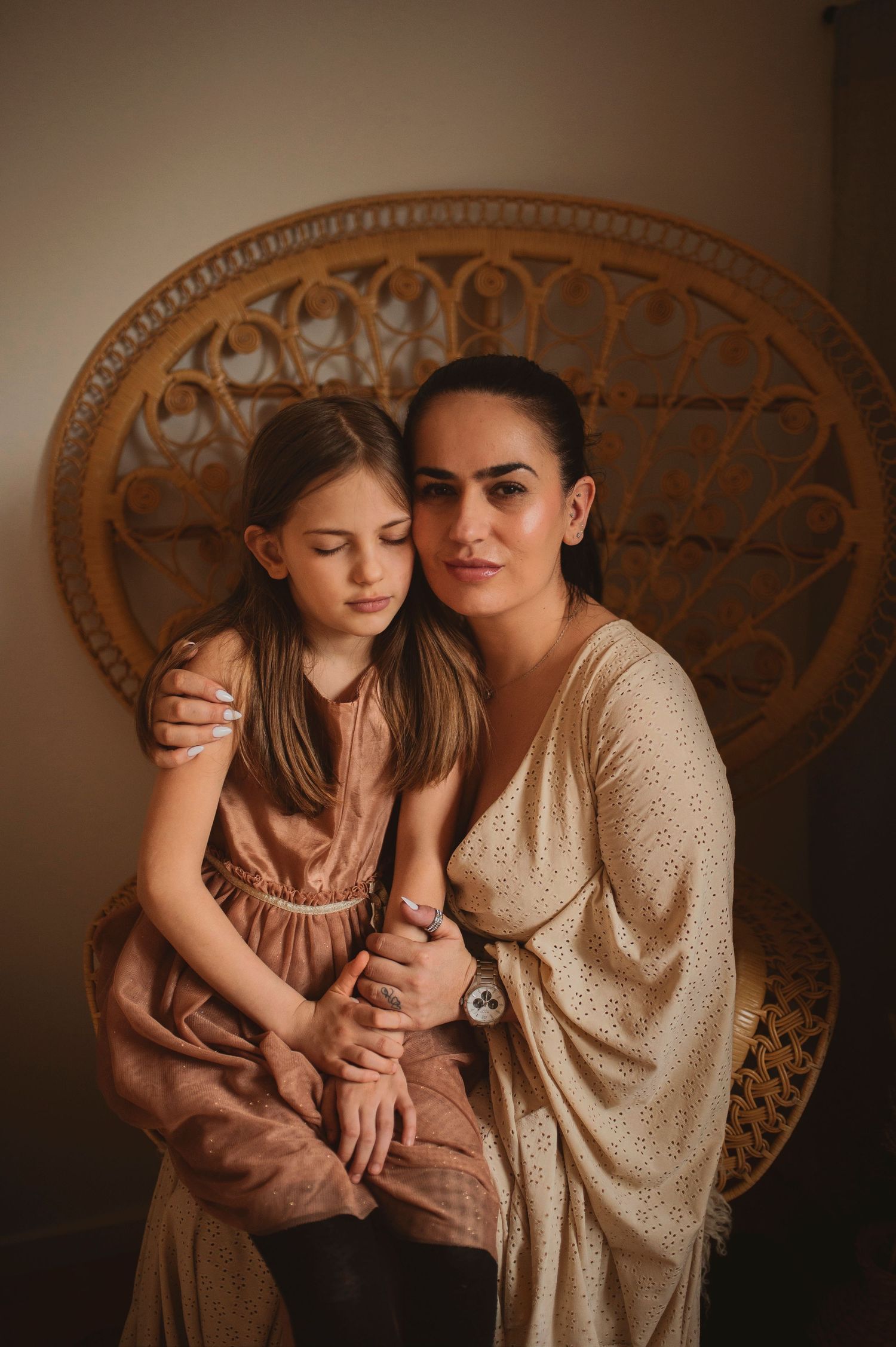 Two figures sit together on a rattan peacock chair in warm earth tones and soft lighting.