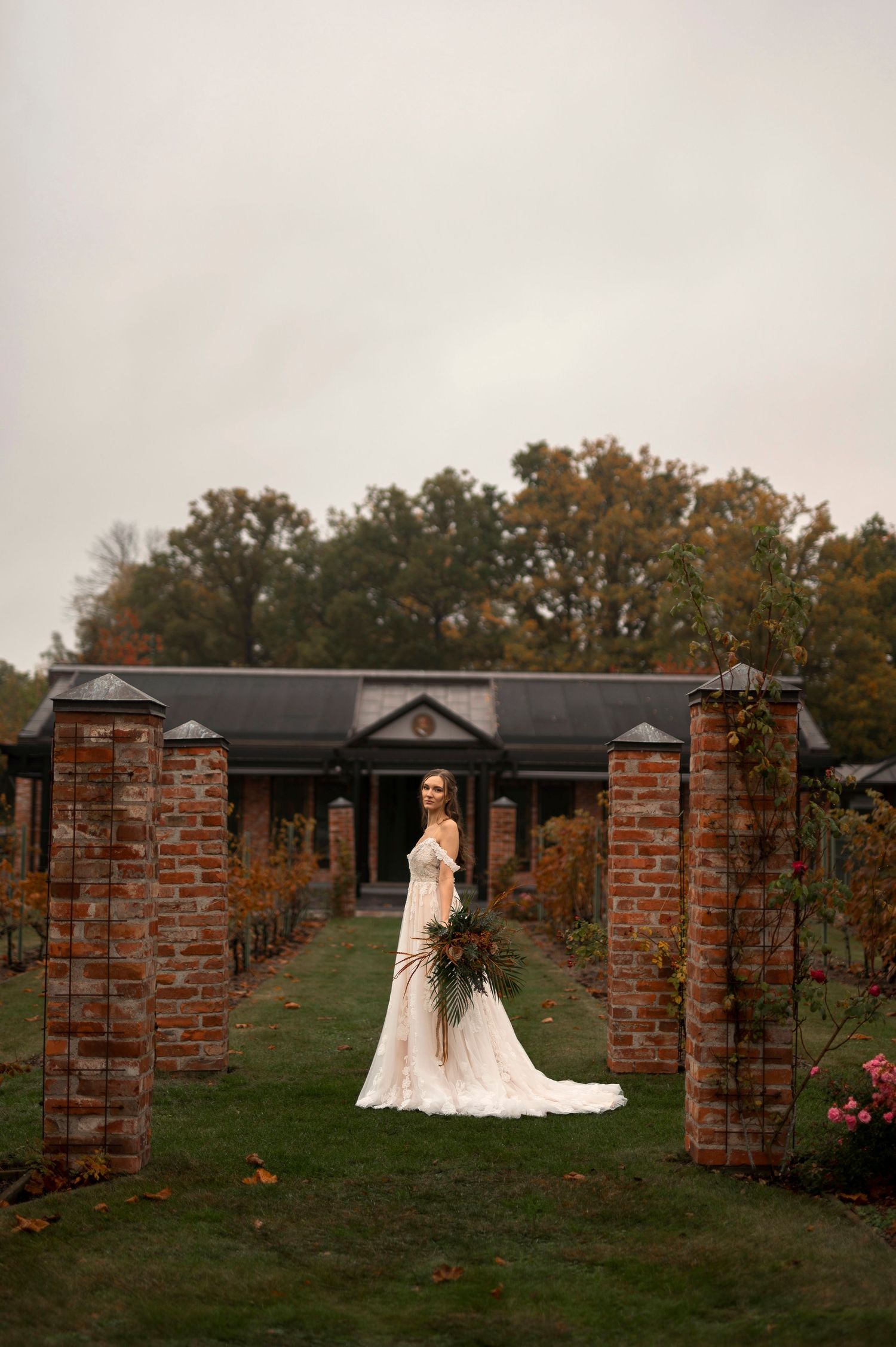 Wedding photography at Åmund Winery  Sweden with bride in white lace gown holding bouquet between brick pillars on an overcast