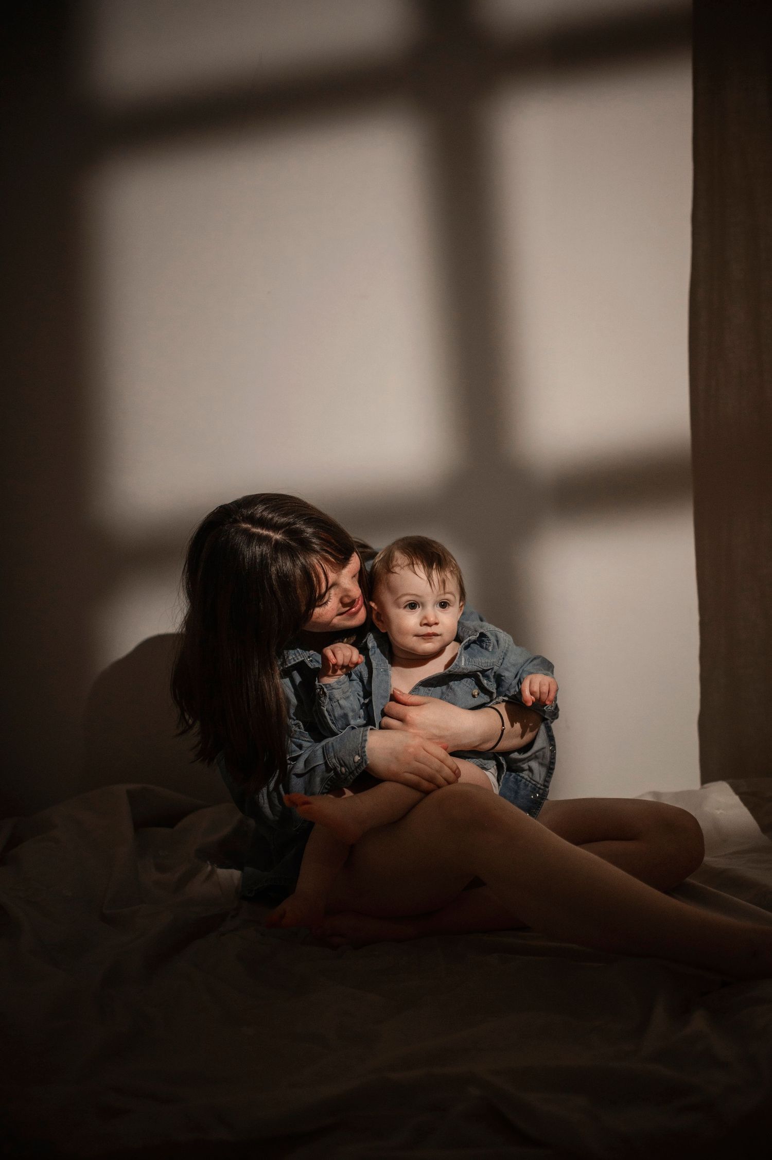 A mother tenderly holds her baby on a bed, bathed in soft natural light from a nearby window.