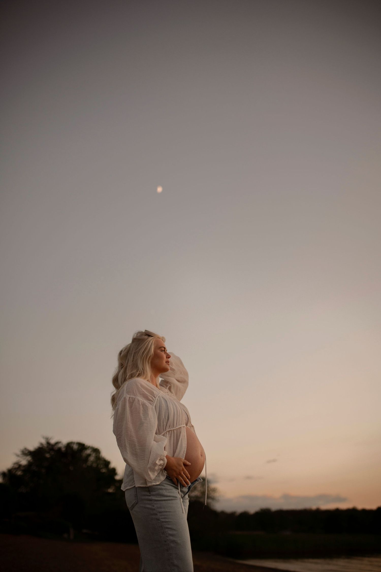 Gravid kvinna fotograferad i solnedgången under en gravidfotografering i Skåne med månen synlig på himlen.