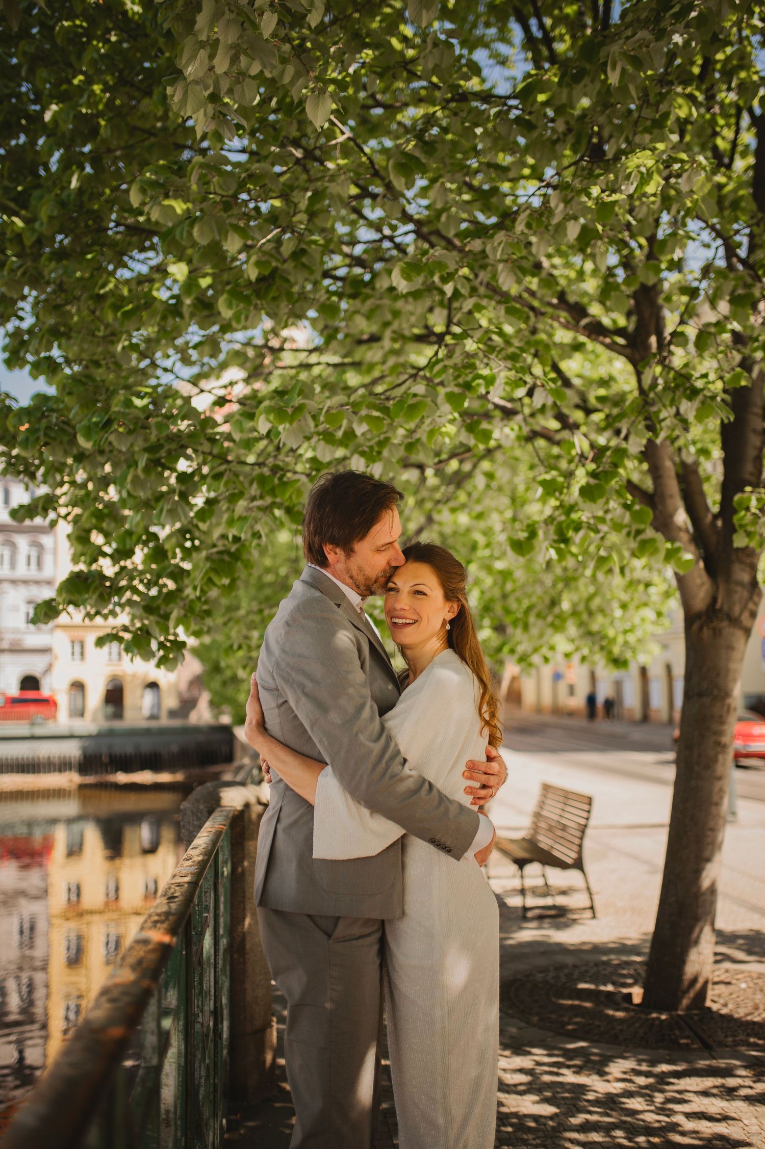 A couple in wedding attire embraces and smiles under green trees by a canal on a sunny day. Elopement in Prauge