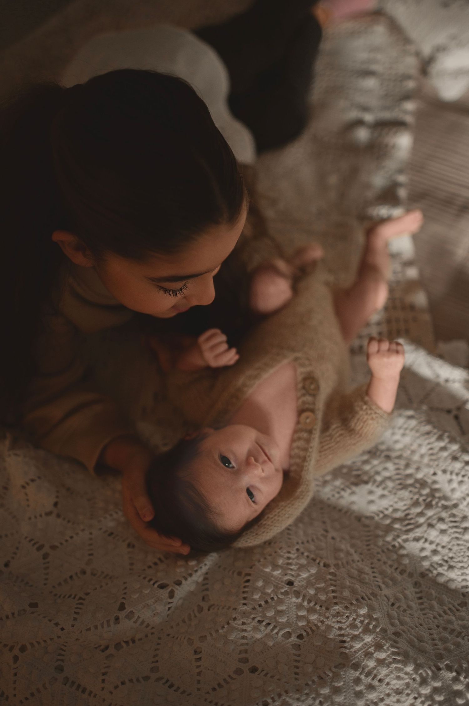 Intimate moment between parent and baby lying on lace blanket in warm lighting.