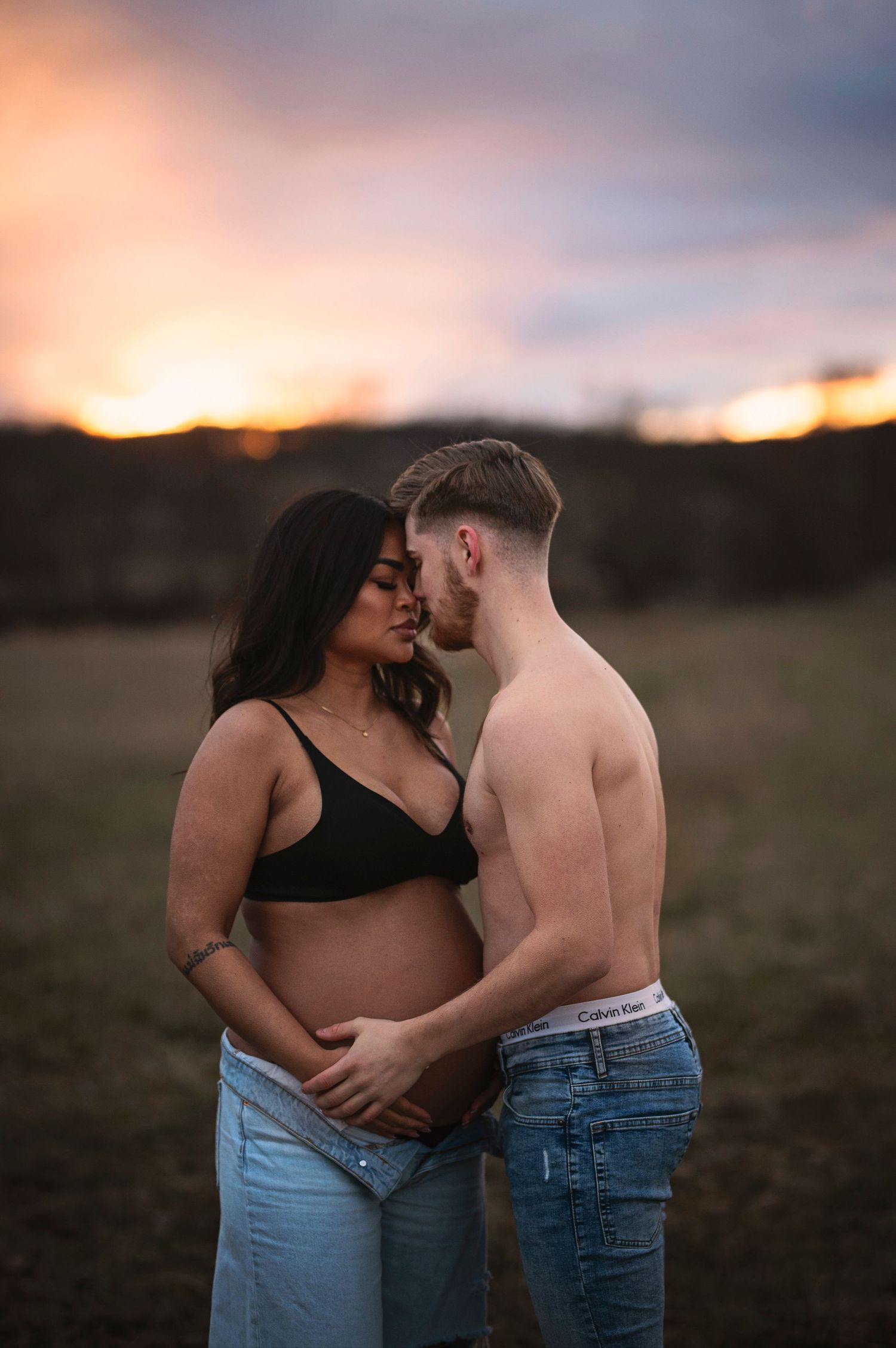 An intimate maternity photo of an expectant couple embracing in a field at golden hour.