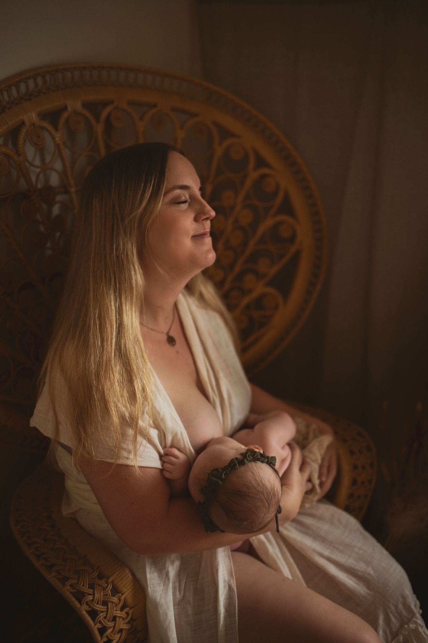 Mother in flowing white dress sits peacefully in vintage chair while nursing in warm natural light.