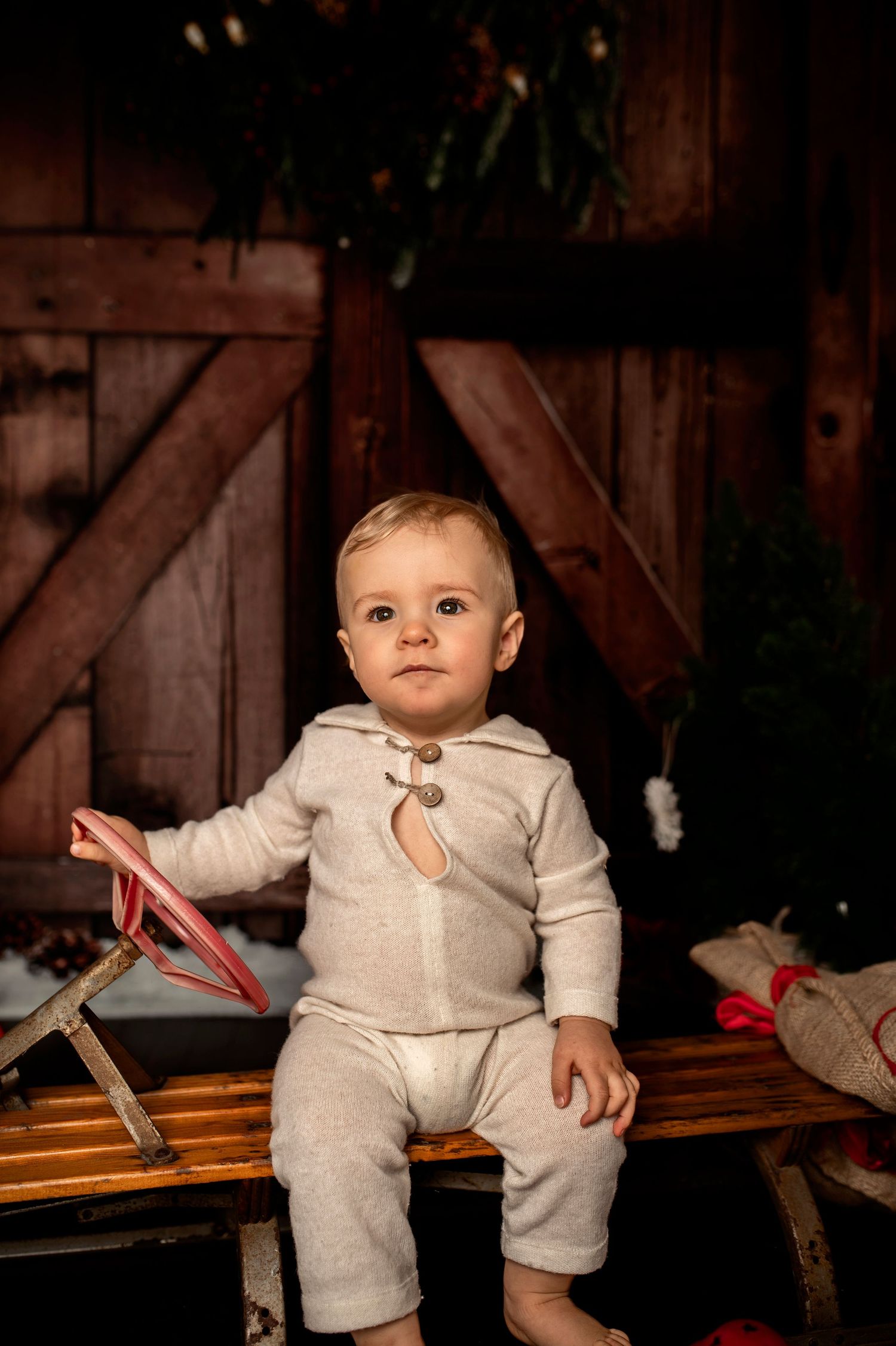 A baby in a cream knit outfit sits on a wooden sled with holiday decor and a rustic barn door backdrop.