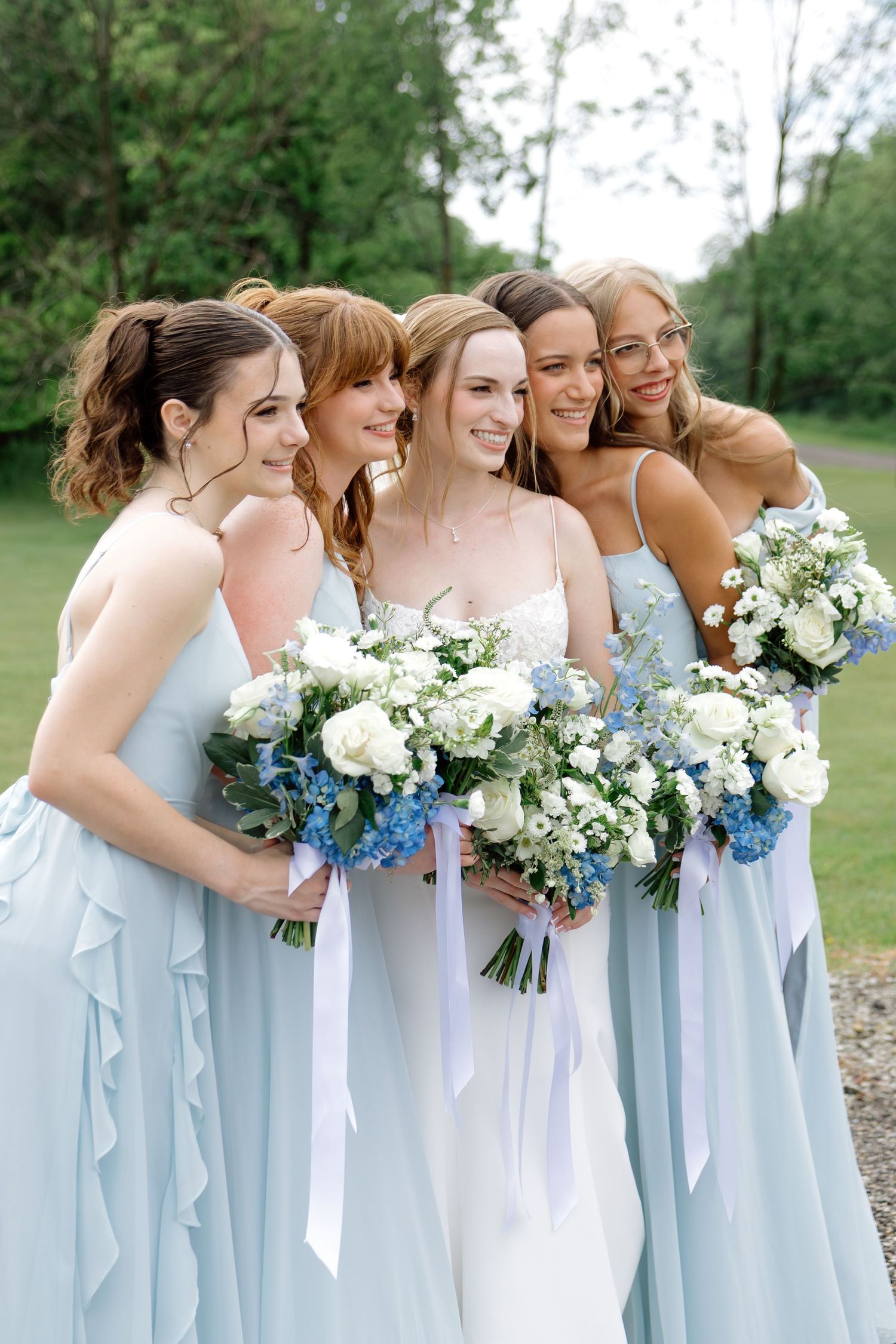 Bride in white gown with four bridesmaids in light blue dresses holding white and blue floral bouquets outdoors.
