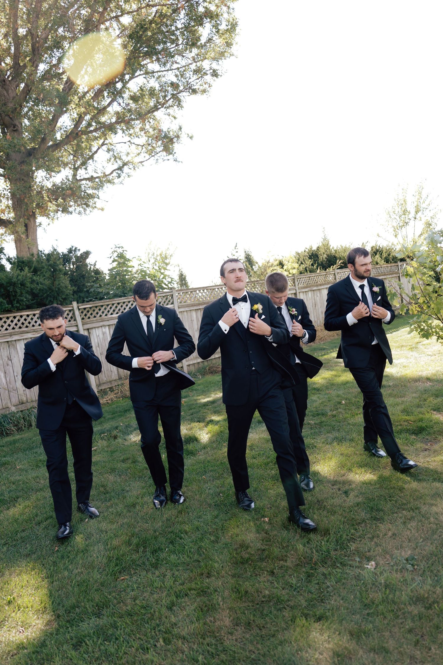 Five groomsmen in black tuxedos adjusting their bow ties on a green lawn during an outdoor wedding.