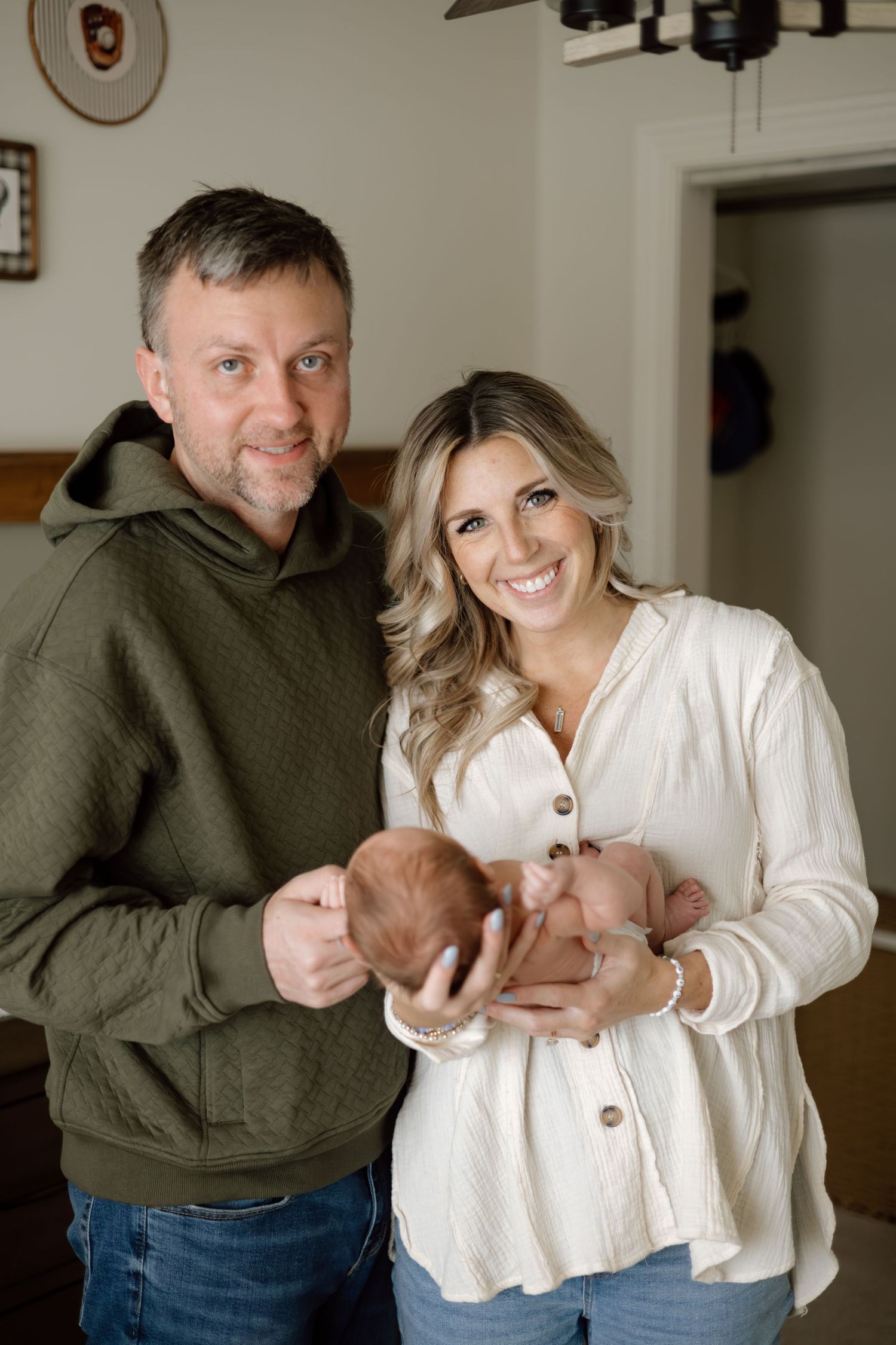 Smiling couple in green and white tops proudly holds their newborn baby in a warm home setting.