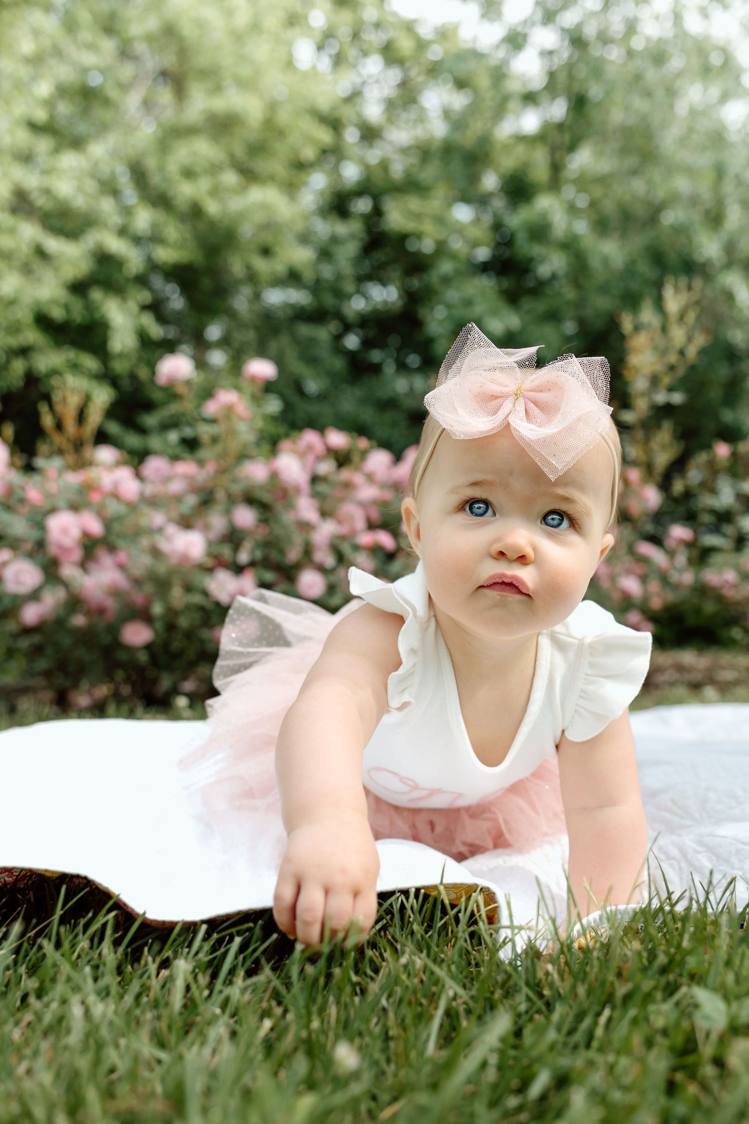 Blue-eyed baby girl in pink tutu and bow headband crawling on grass in a garden.