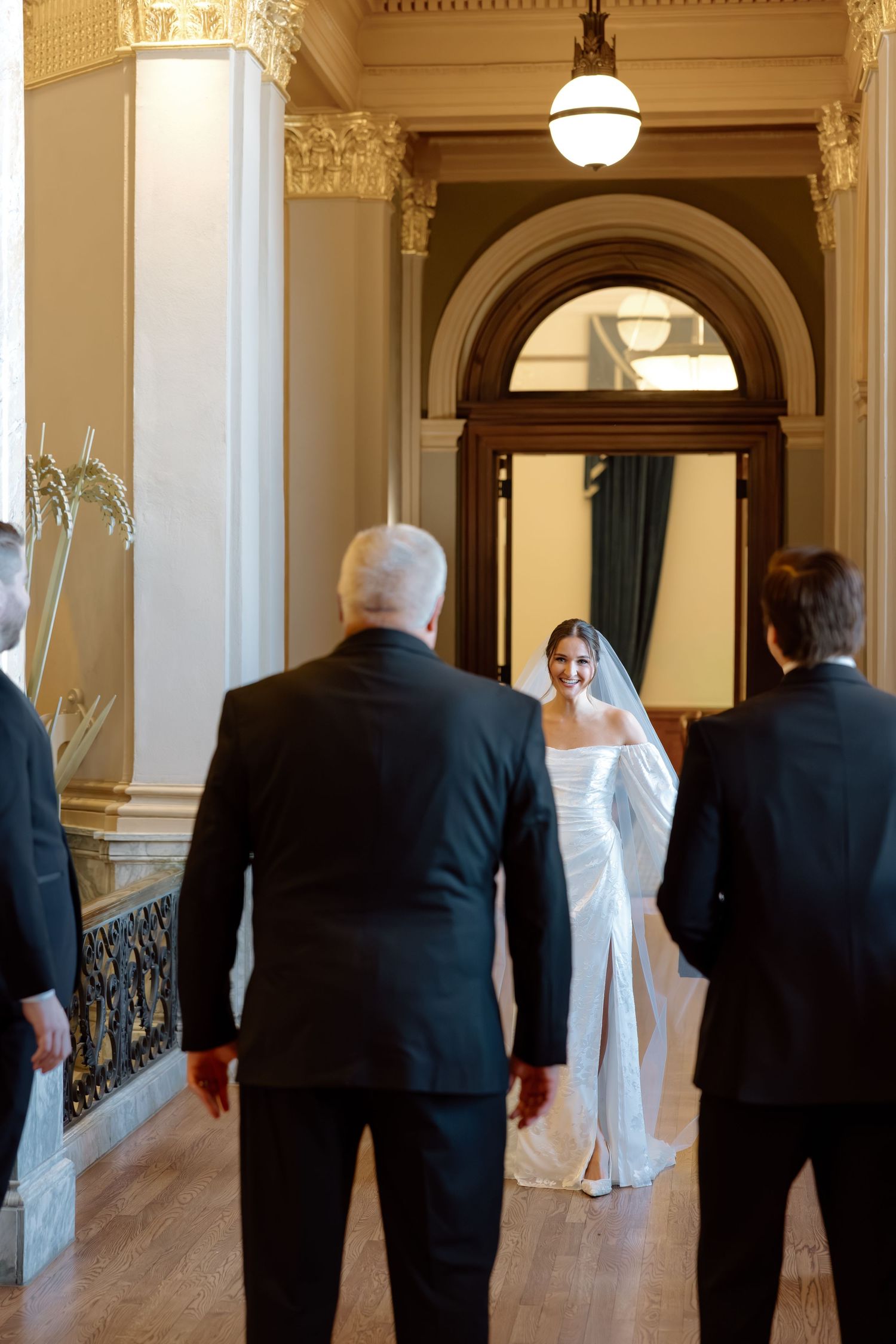 Bride in white gown and veil walks toward groom and groomsmen in grand ornate wedding venue hallway.