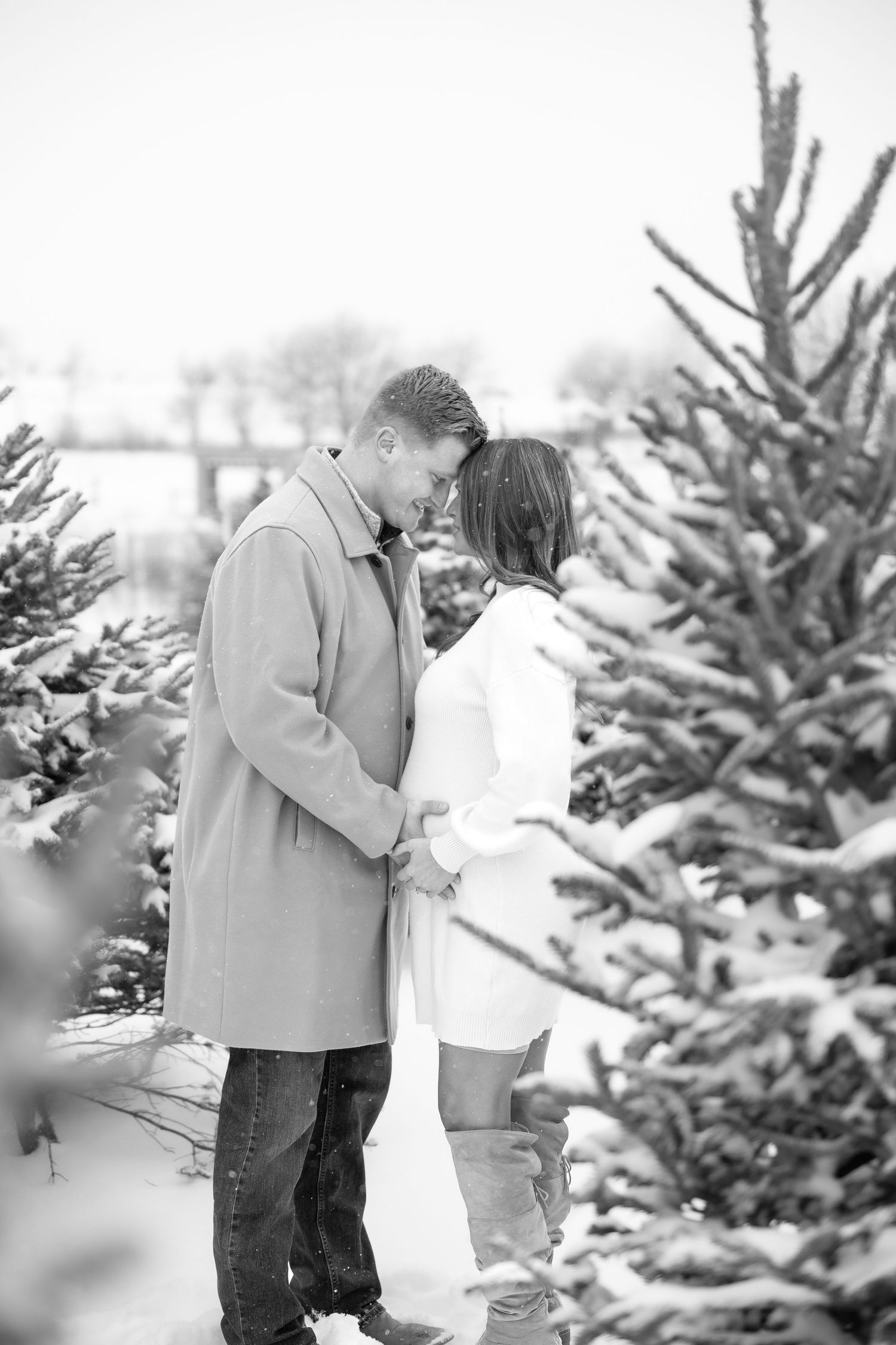 Black and white photo of expecting couple holding hands among snow-dusted Christmas trees.