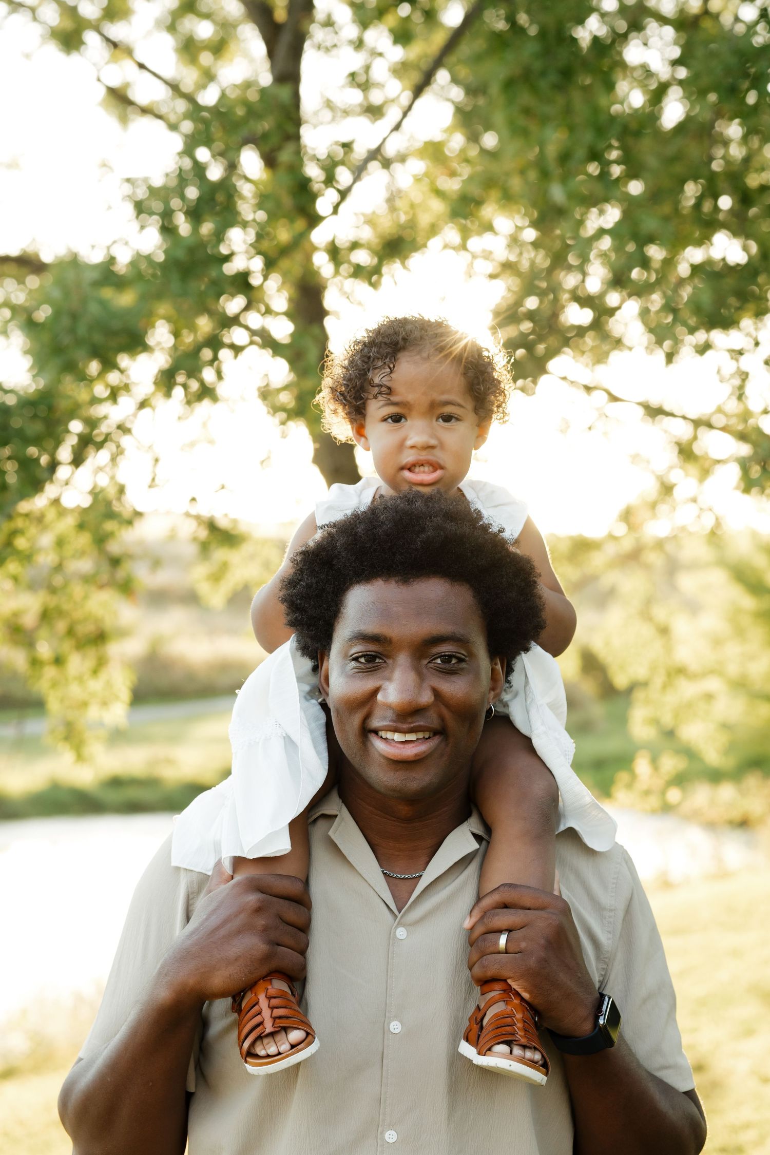 Smiling father carrying toddler daughter on his shoulders in a park.
