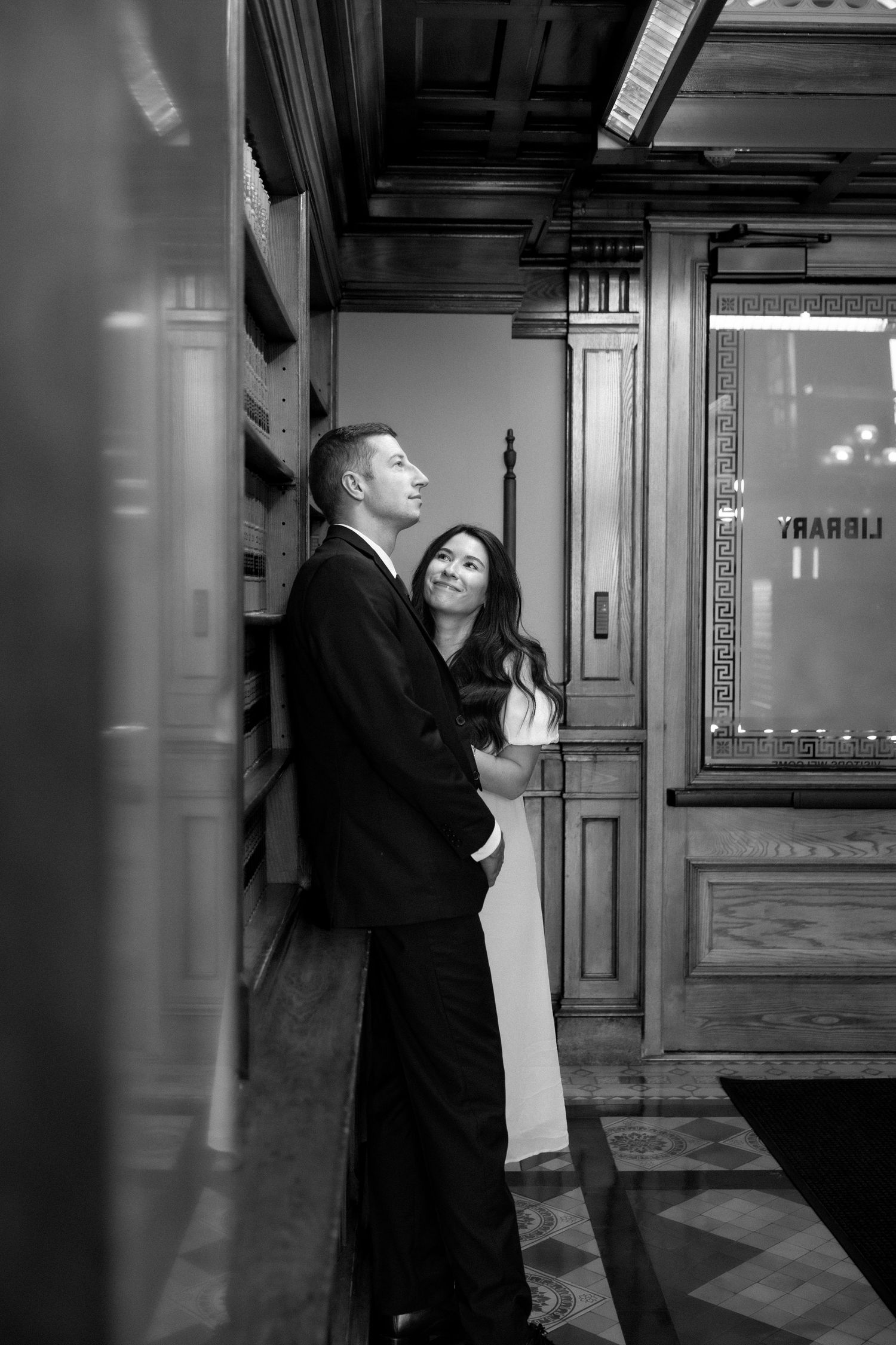 Black and white photo of couple embracing in a historic wood-paneled library corridor.
