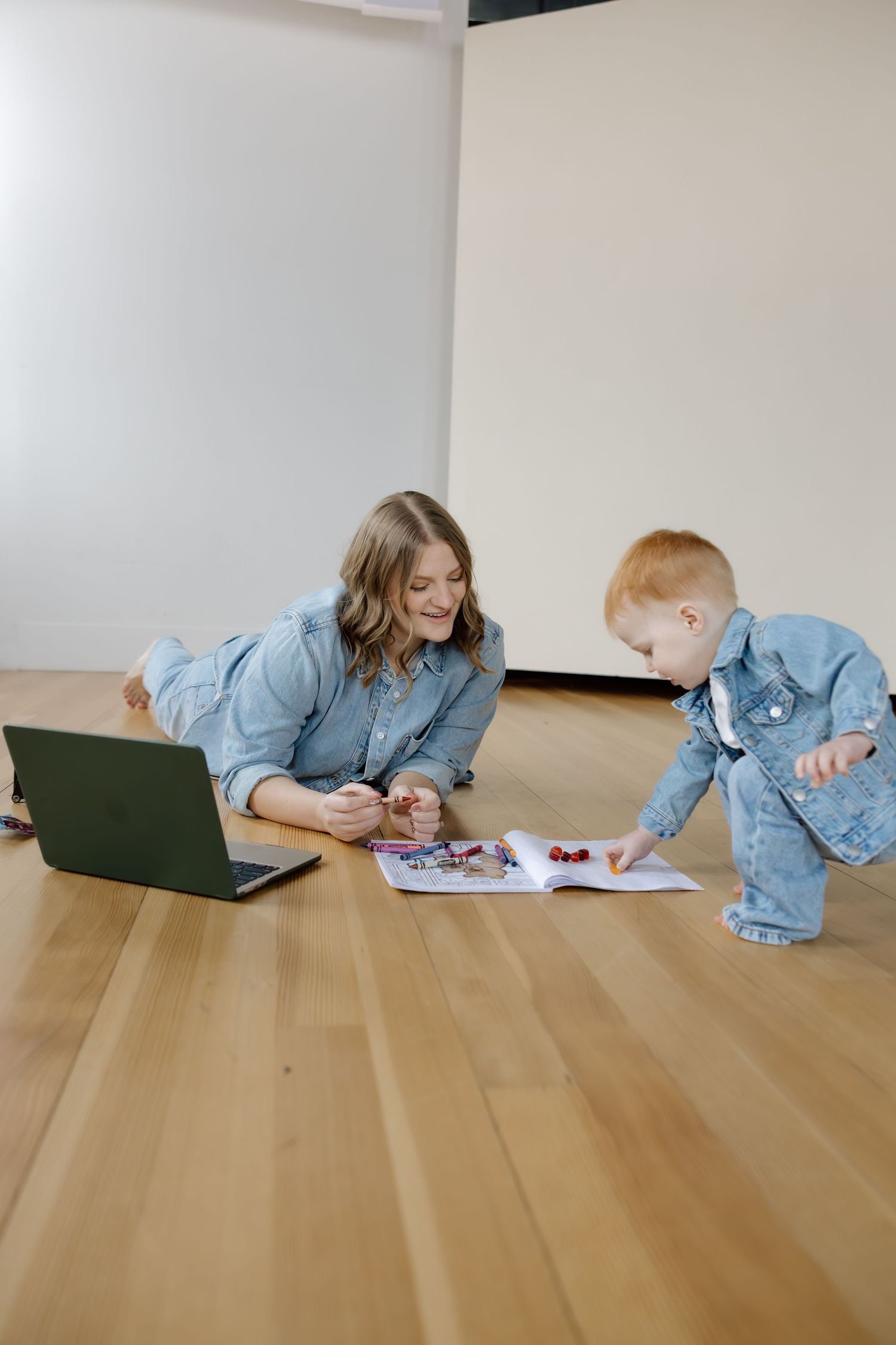 Mother in denim shirt smiles at toddler looking at colorful book on hardwood floor with laptop nearby.