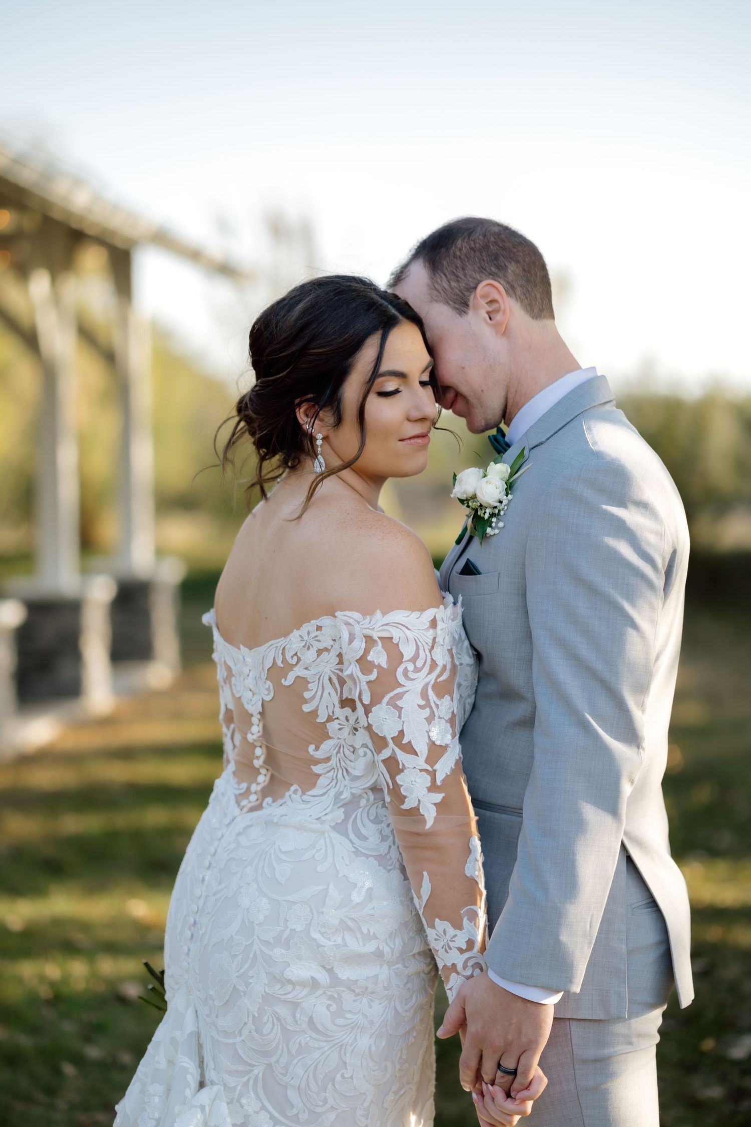 Bride in lace off-shoulder gown and groom in gray suit share tender moment outdoors at golden hour wedding.
