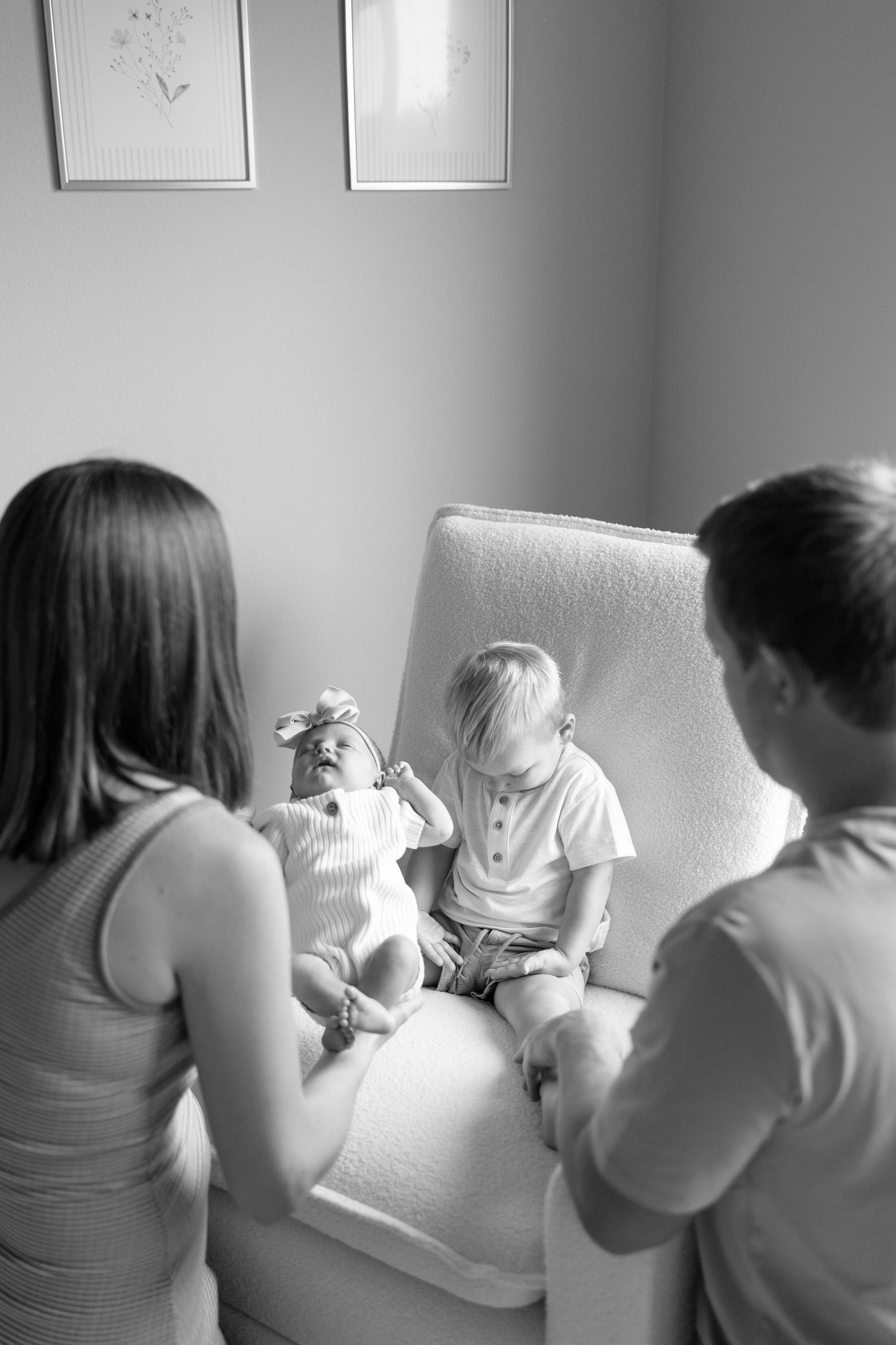 Black and white photo of parents watching their two young children sit together on a white armchair.