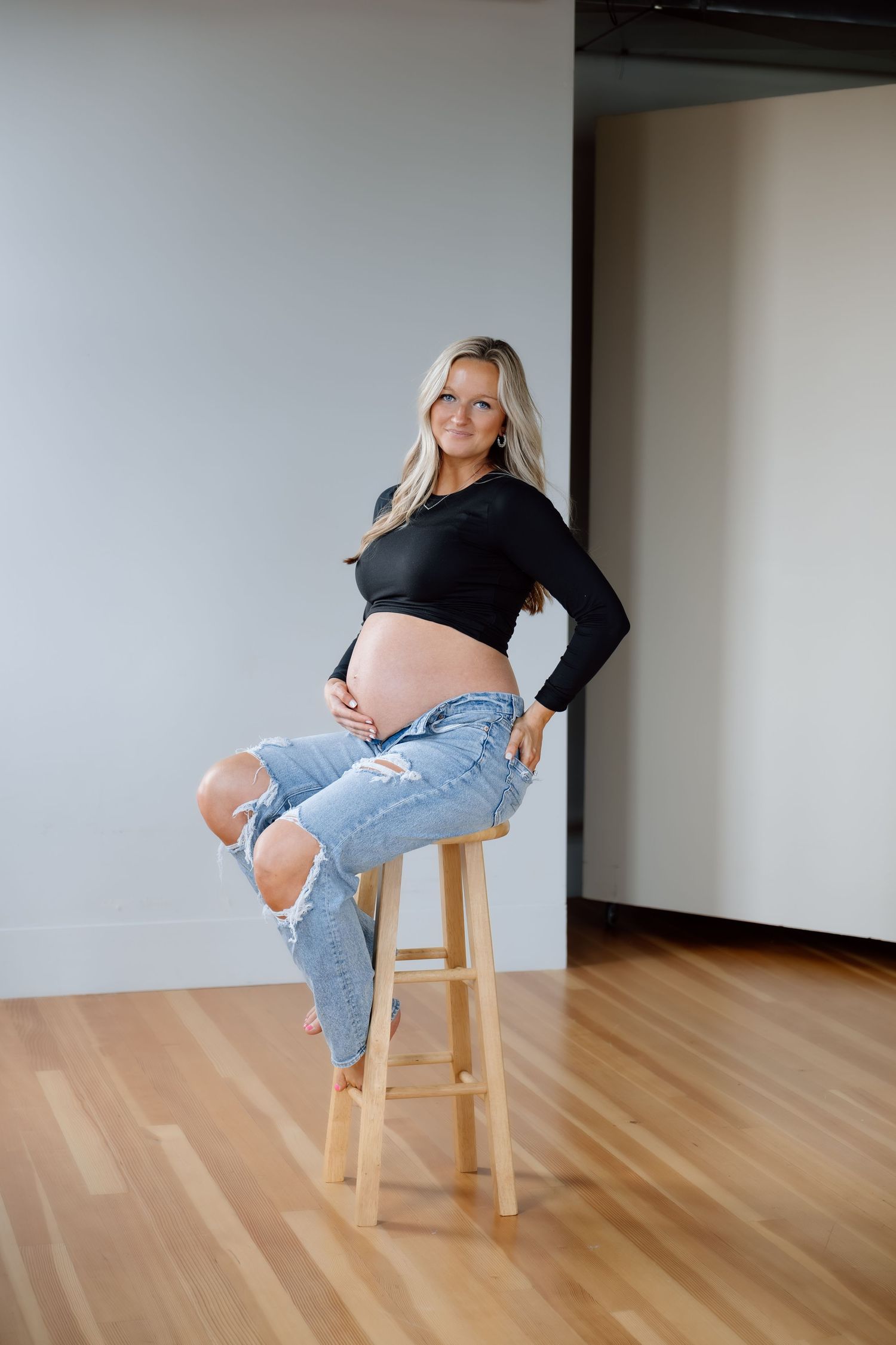 Pregnant woman in black crop top and ripped jeans sitting on wooden stool showing baby bump.