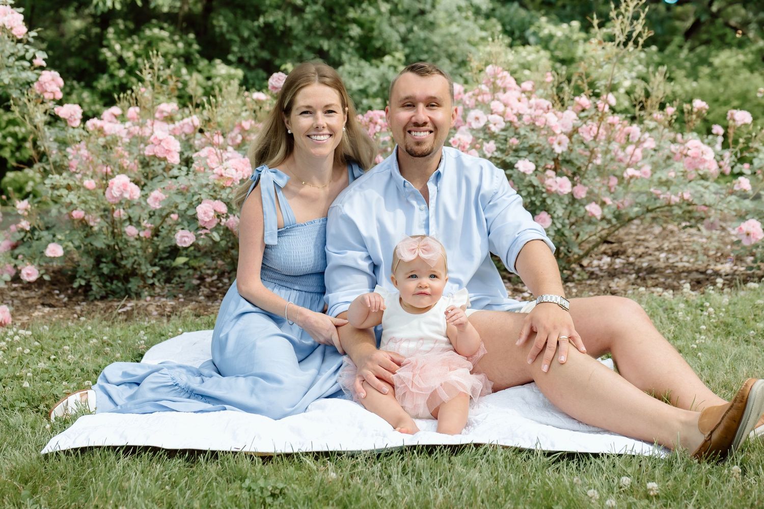 Happy family of three sits on blanket in garden with pink roses, parents in matching blue outfits with baby.