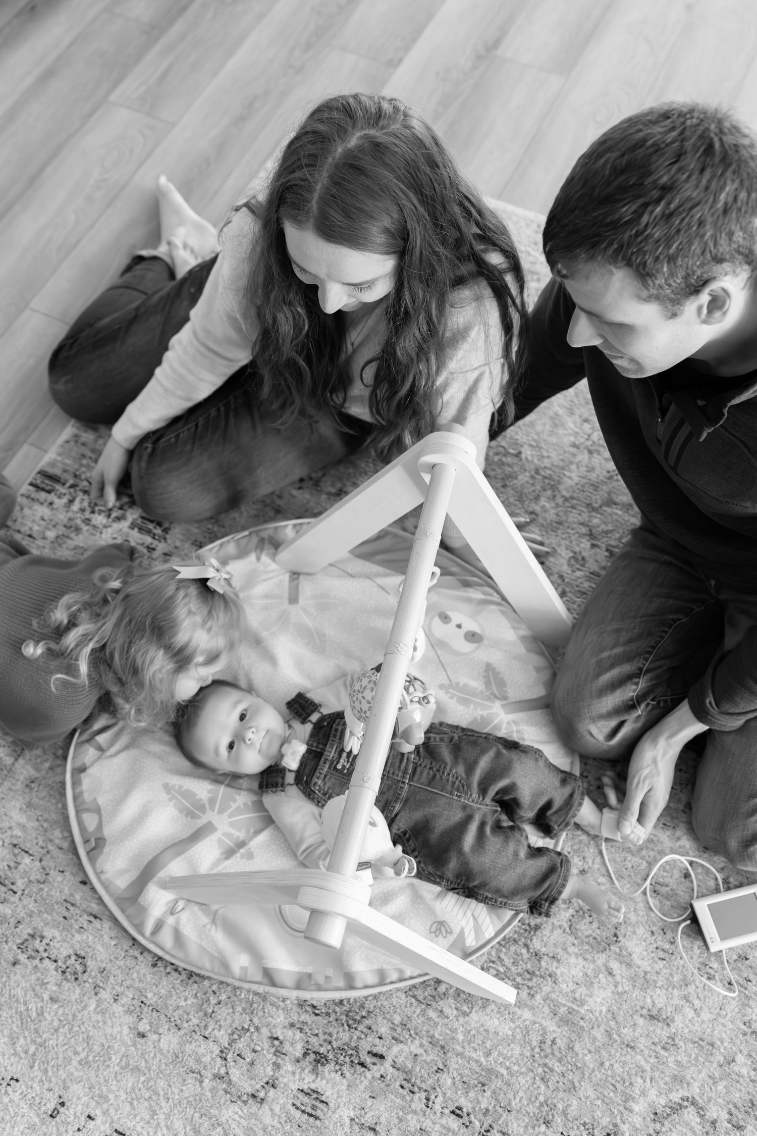 Black and white overhead photo of parents sitting on floor watching baby under play gym at home. Des Moines Newborn Photographer.