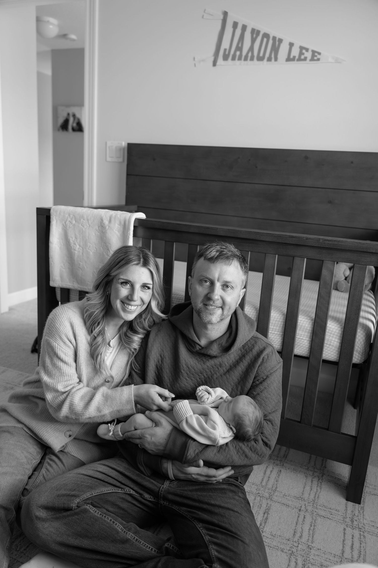 Black and white photo of parents sitting with newborn in nursery below Jaxon Lee name banner.