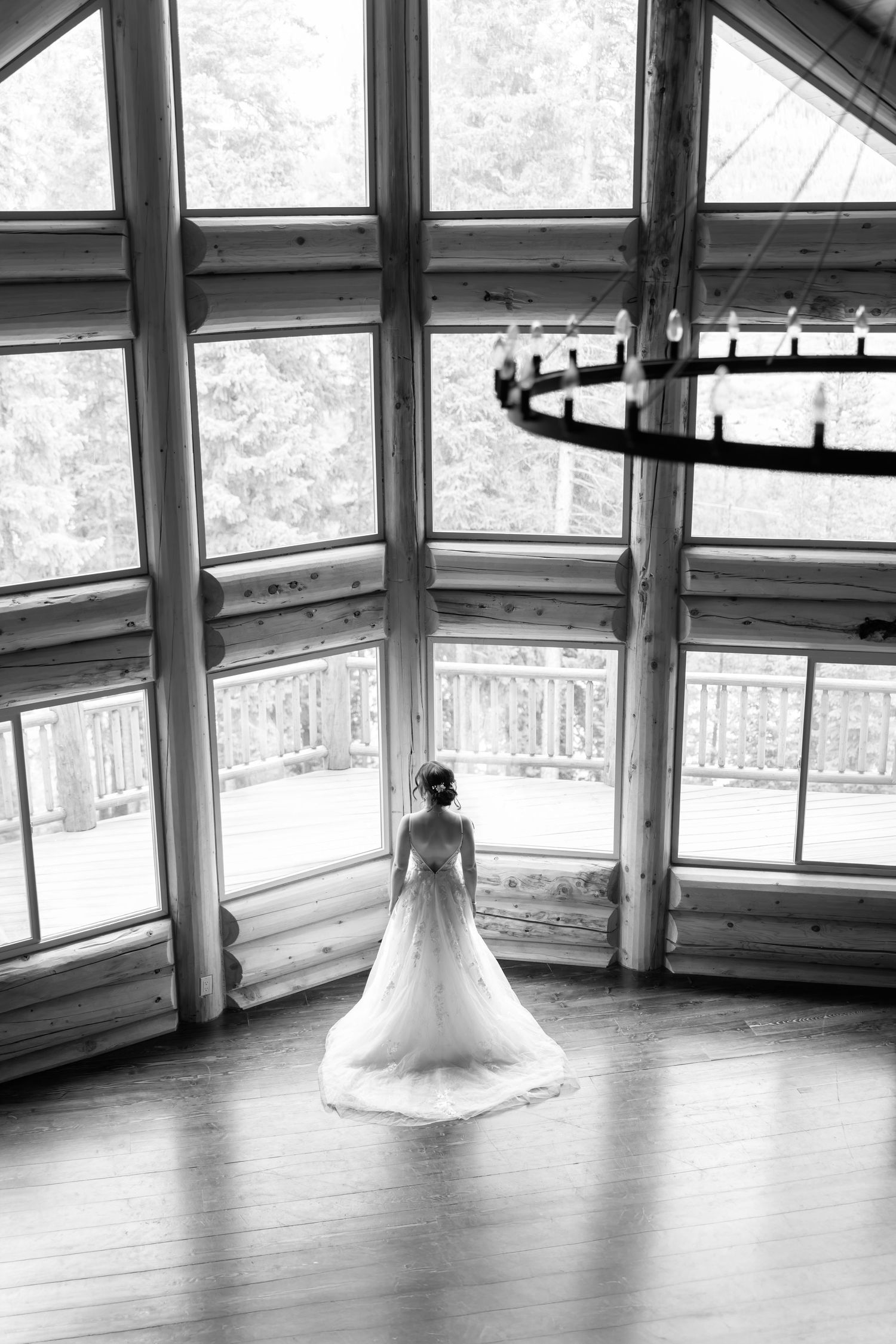 Bride in a flowing white wedding gown stands before large windows in a rustic log cabin venue, black and white photo.