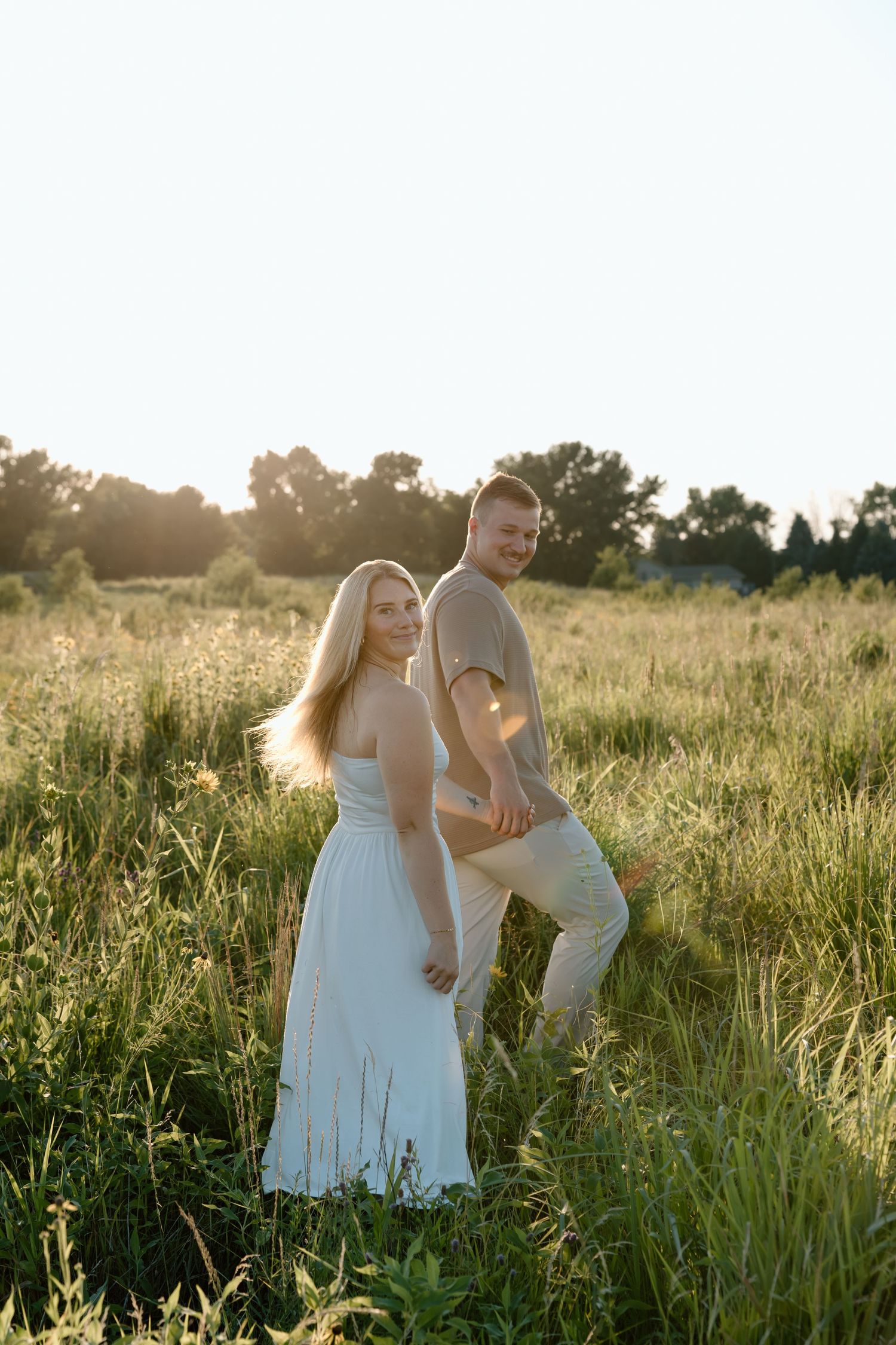 A couple stands in a sunlit green field, the woman in a white dress smiling warmly.