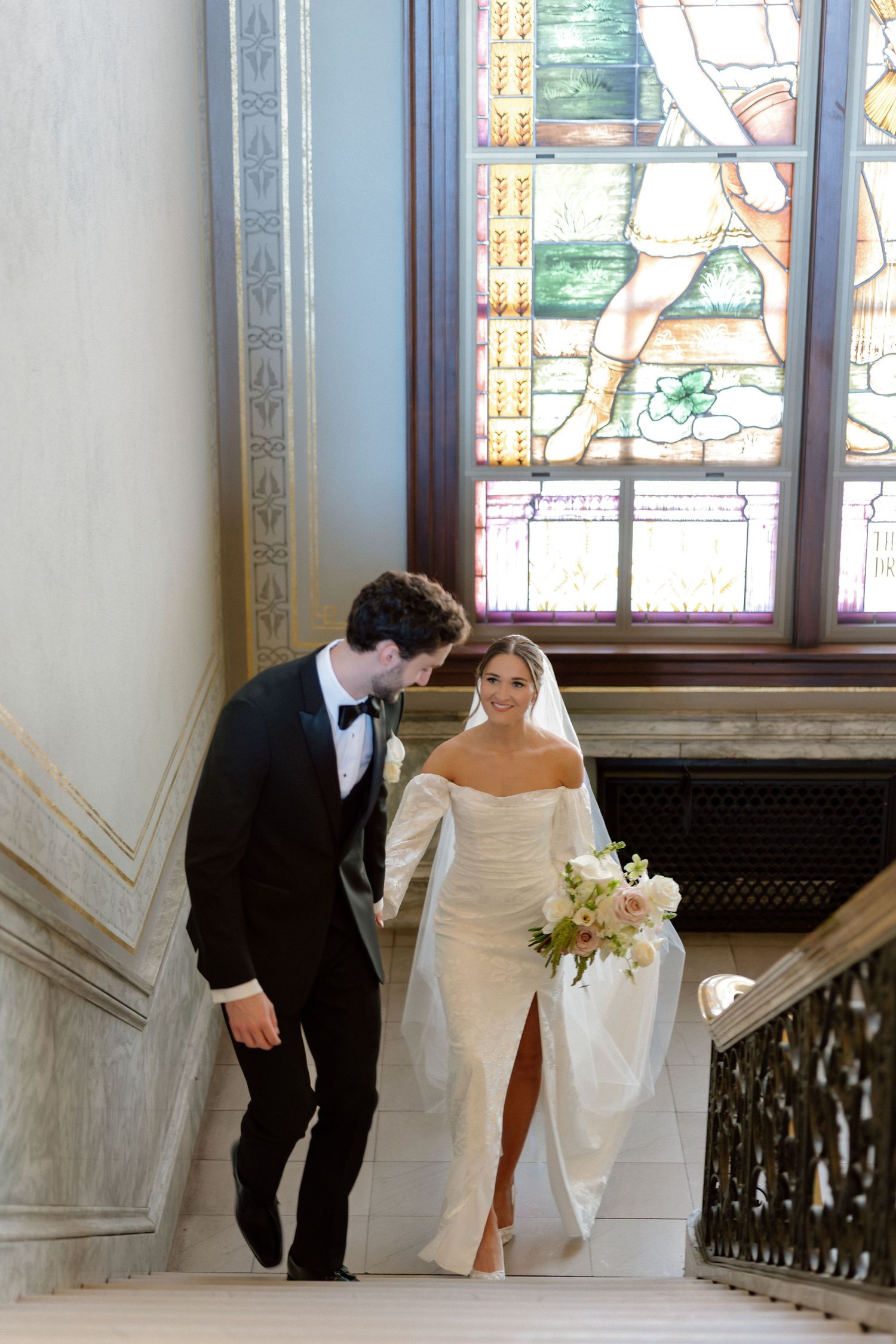 Bride and groom walk up grand staircase with stained glass window, bride in off-shoulder gown holding bouquet.