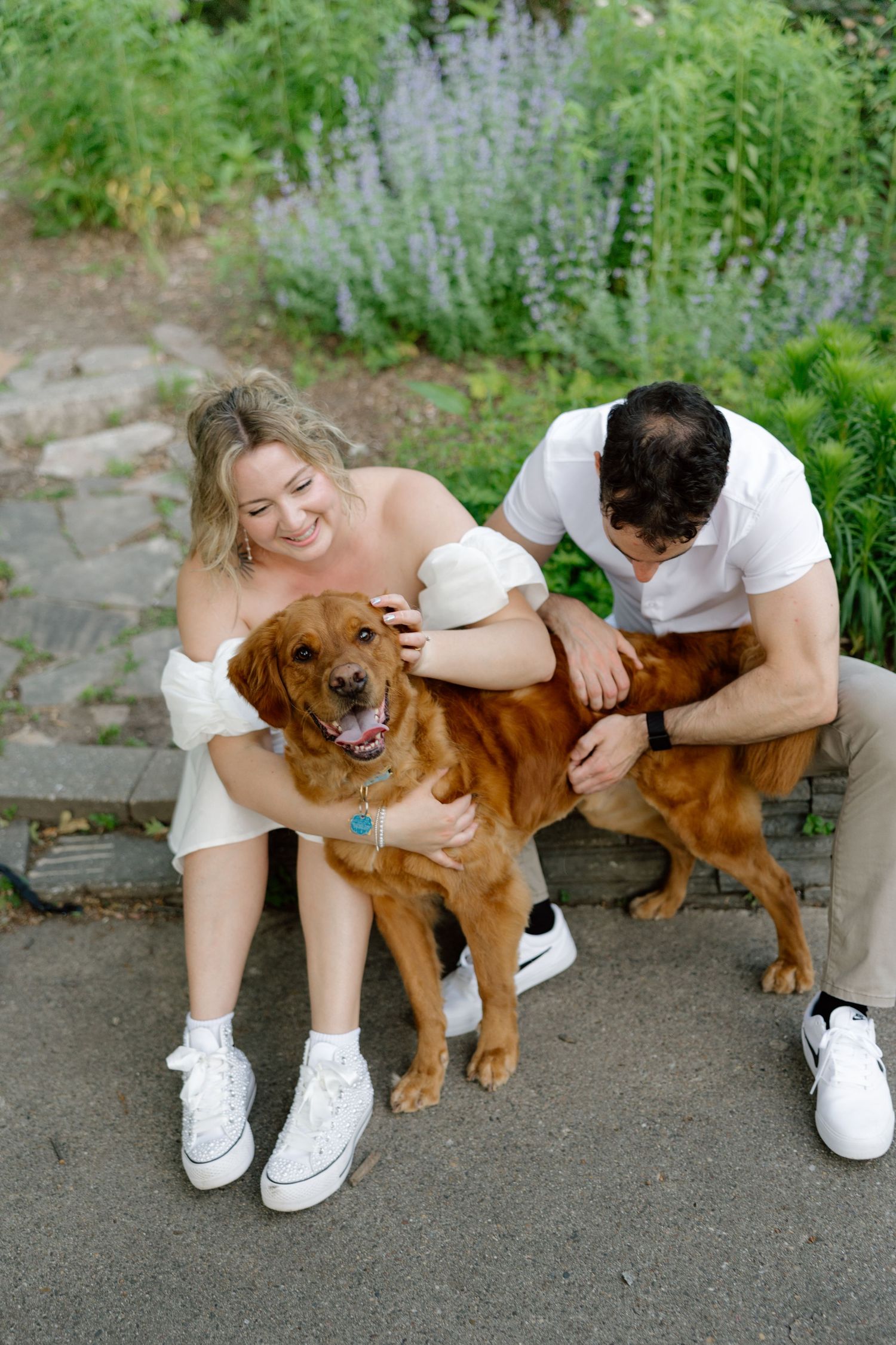 A smiling couple sits outdoors petting their golden retriever dog, surrounded by lush green garden foliage.