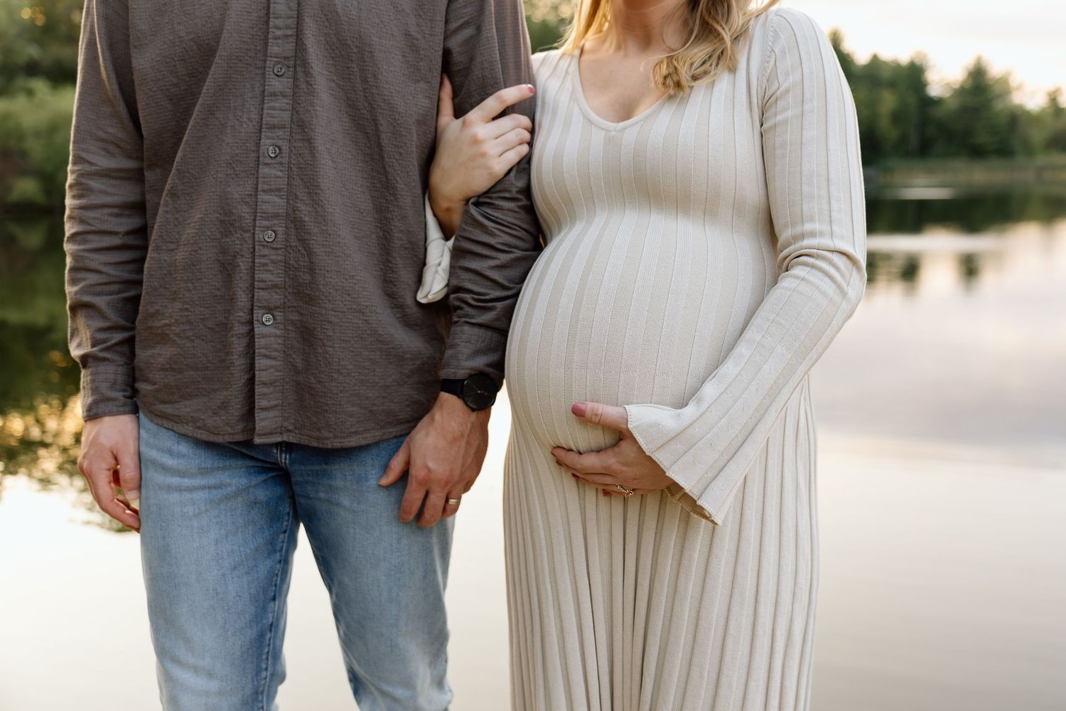 Close-up of pregnant woman in white ribbed dress holding baby bump beside partner near reflective lake.