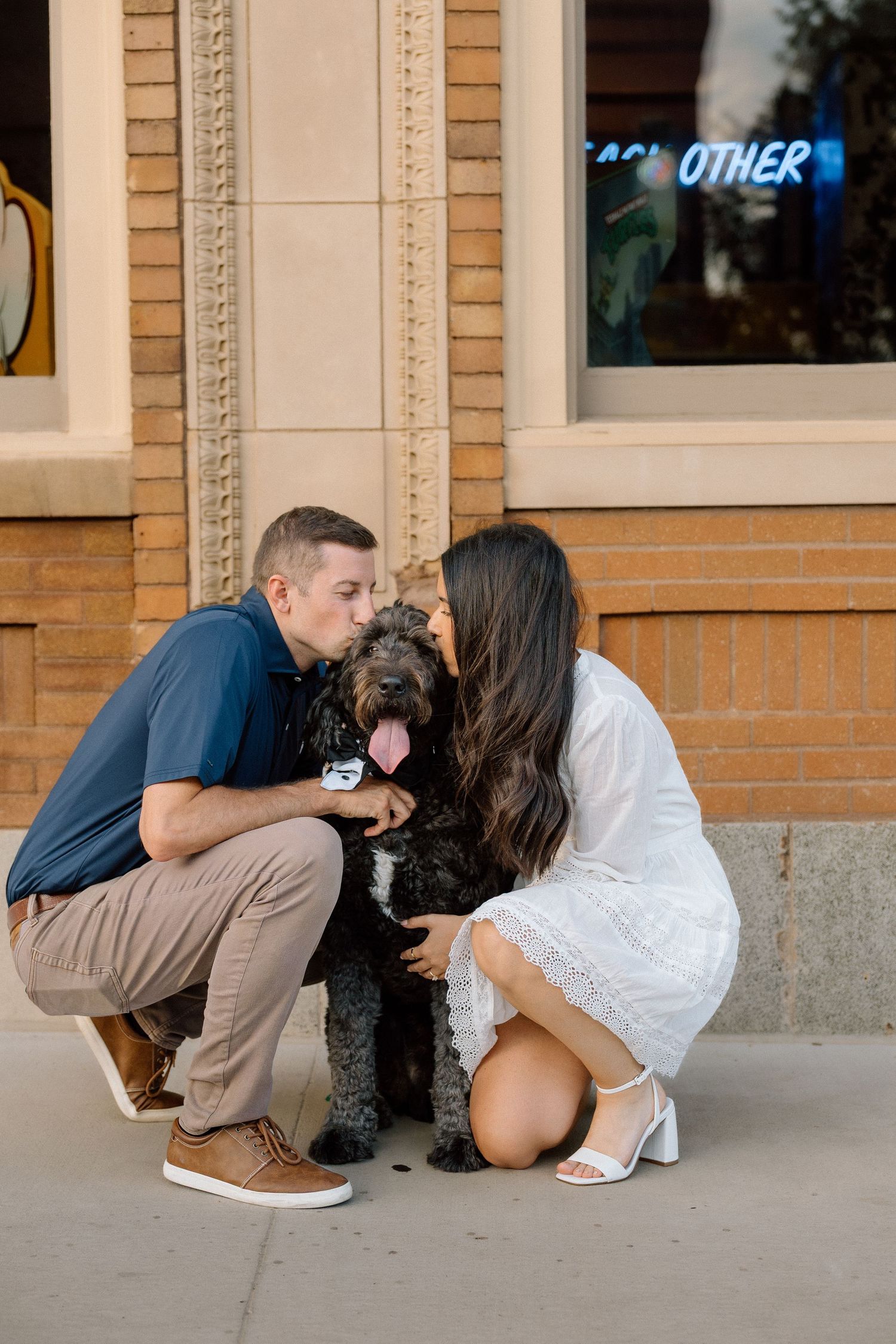 Couple crouching down to kiss their black doodle dog on the cheek outside a brick storefront.