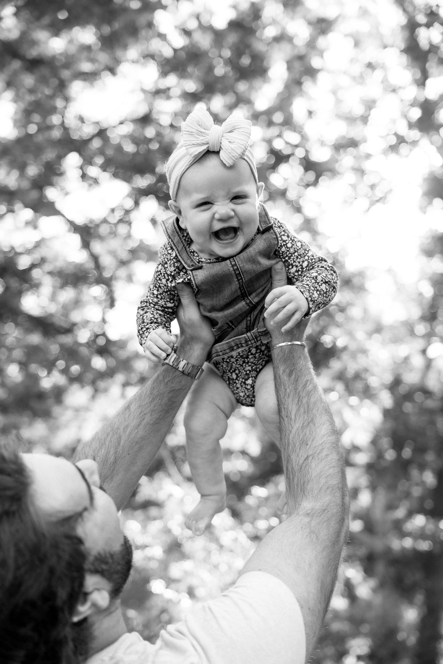 Black and white photo of joyful baby girl in bow headband lifted high by fathers hands.