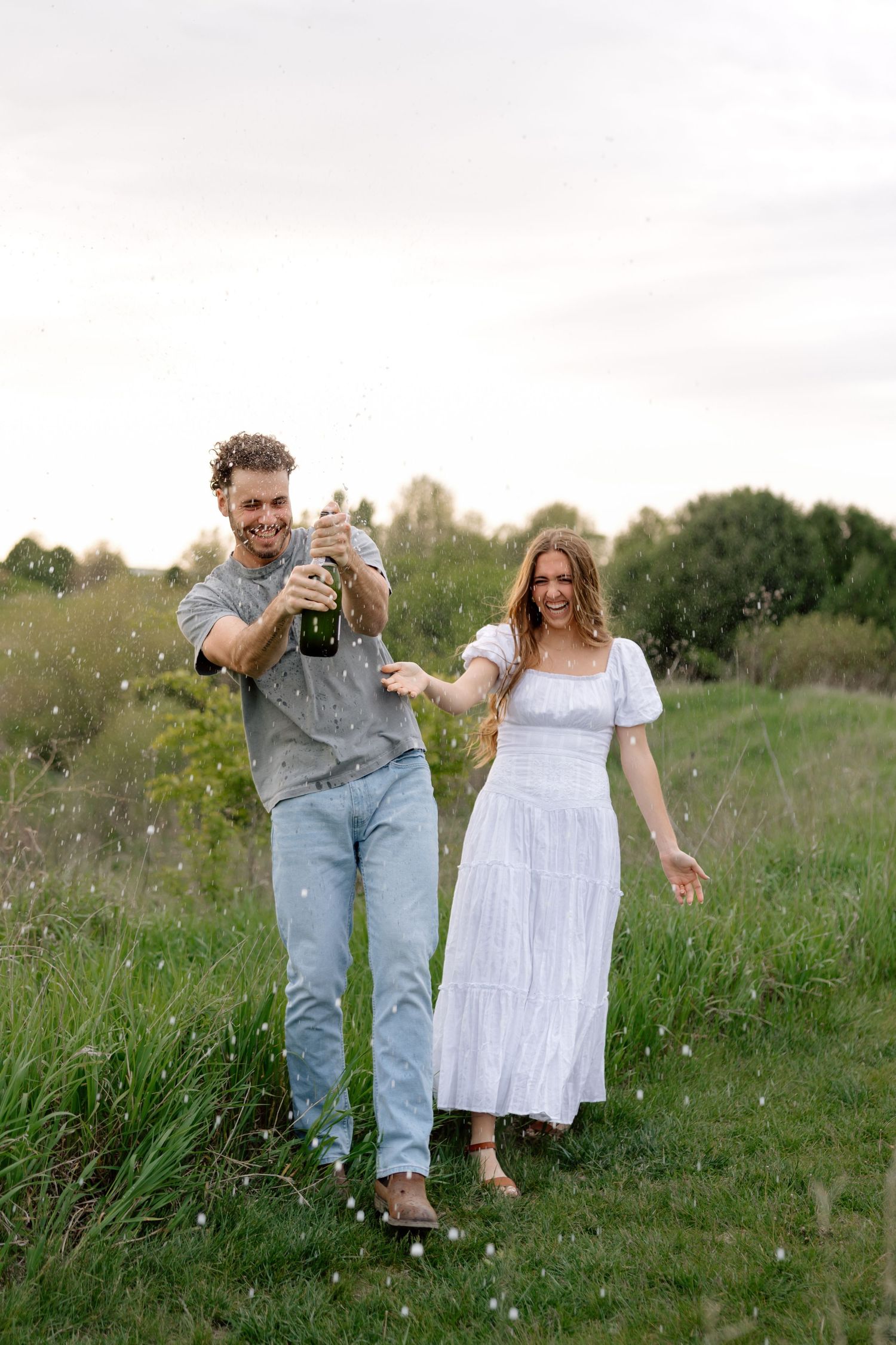Joyful couple celebrating outdoors, man popping champagne bottle while woman in white dress laughs in green meadow.