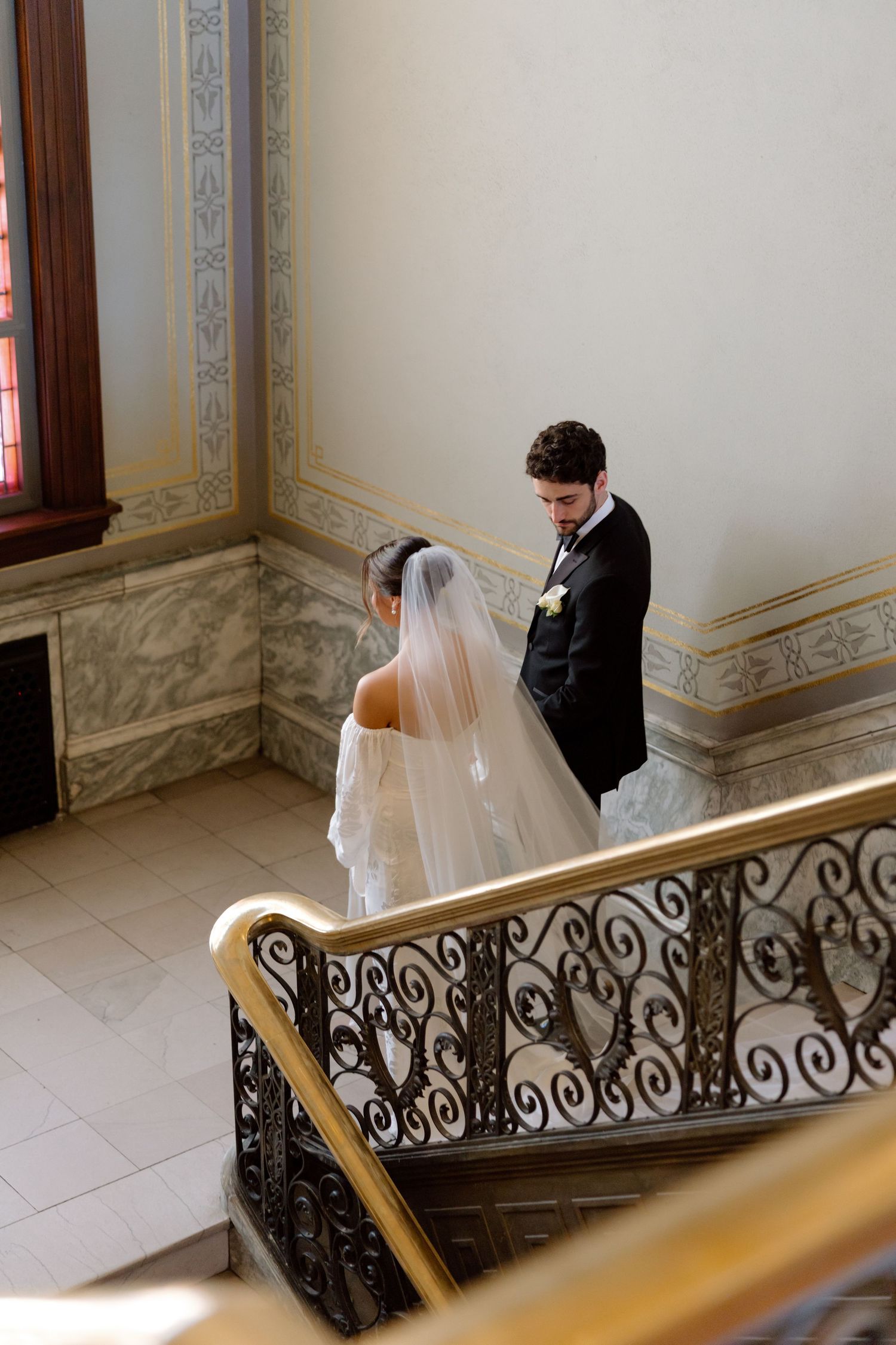 Bride and groom descend an ornate marble staircase with iron railings in an elegant wedding venue.