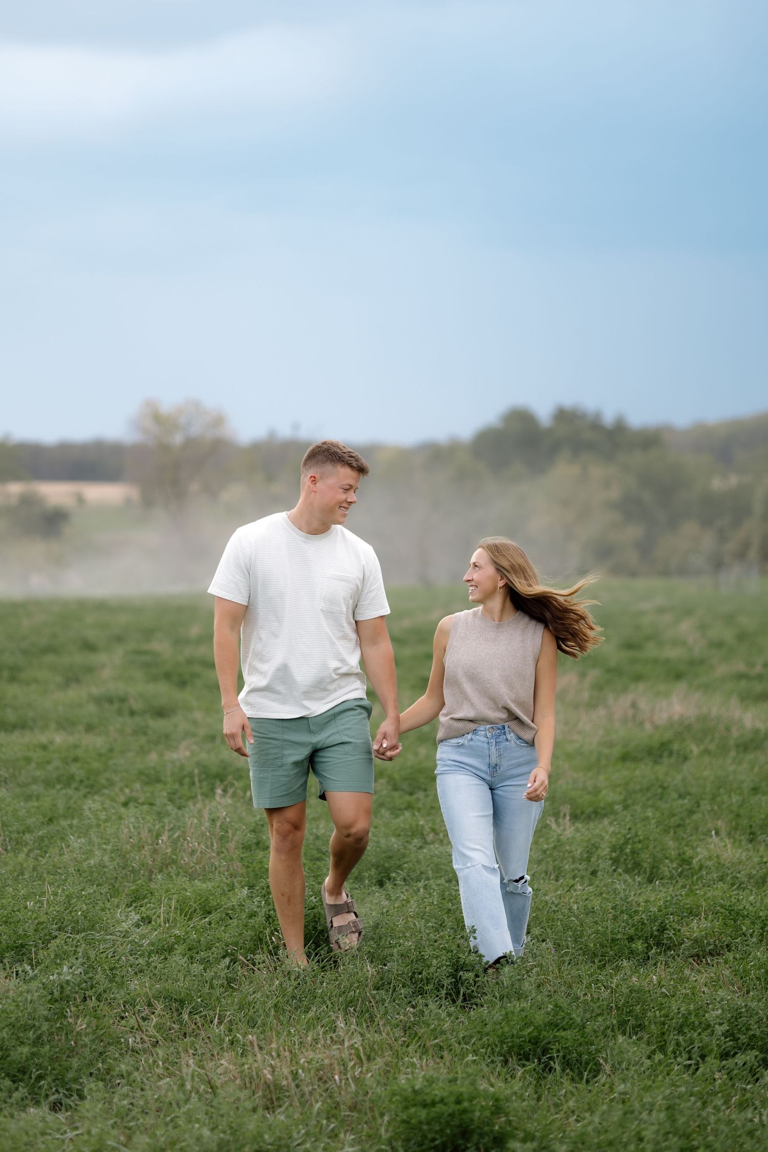 Young couple holding hands walking through green field with dramatic cloudy sky in background.