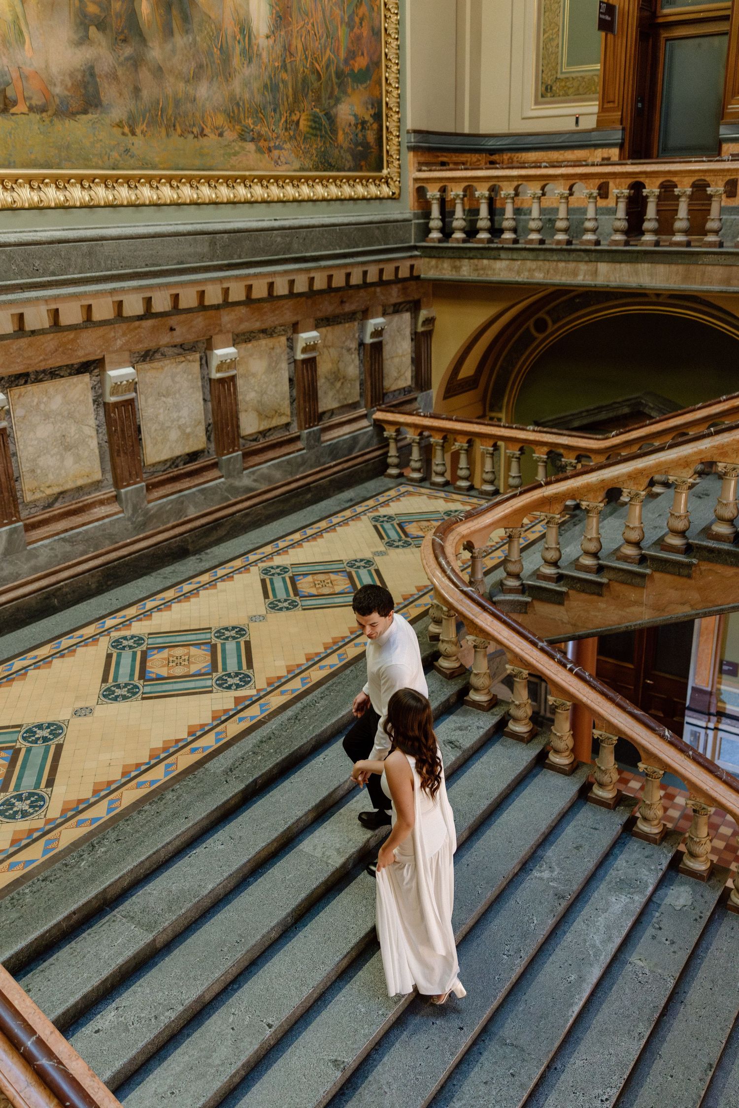 Couple in wedding attire descends grand ornate staircase inside a stunning historic capitol building.